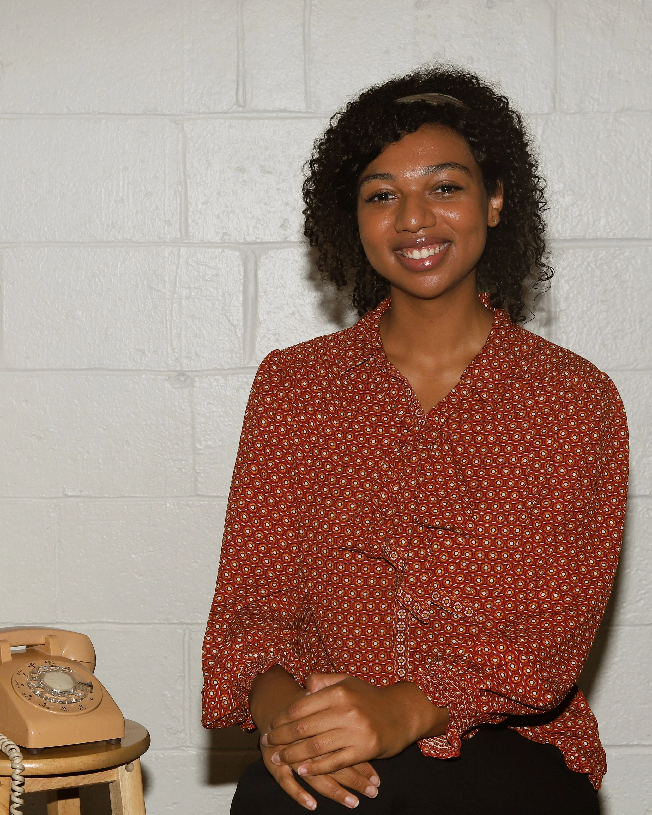 Kendelle, a black woman, is sitting smiling at the camera, with her hands in her lap. She wears an orange patterned blouse. To her left, is a stool with an old rotary phone sitting on it. The background is white brick.