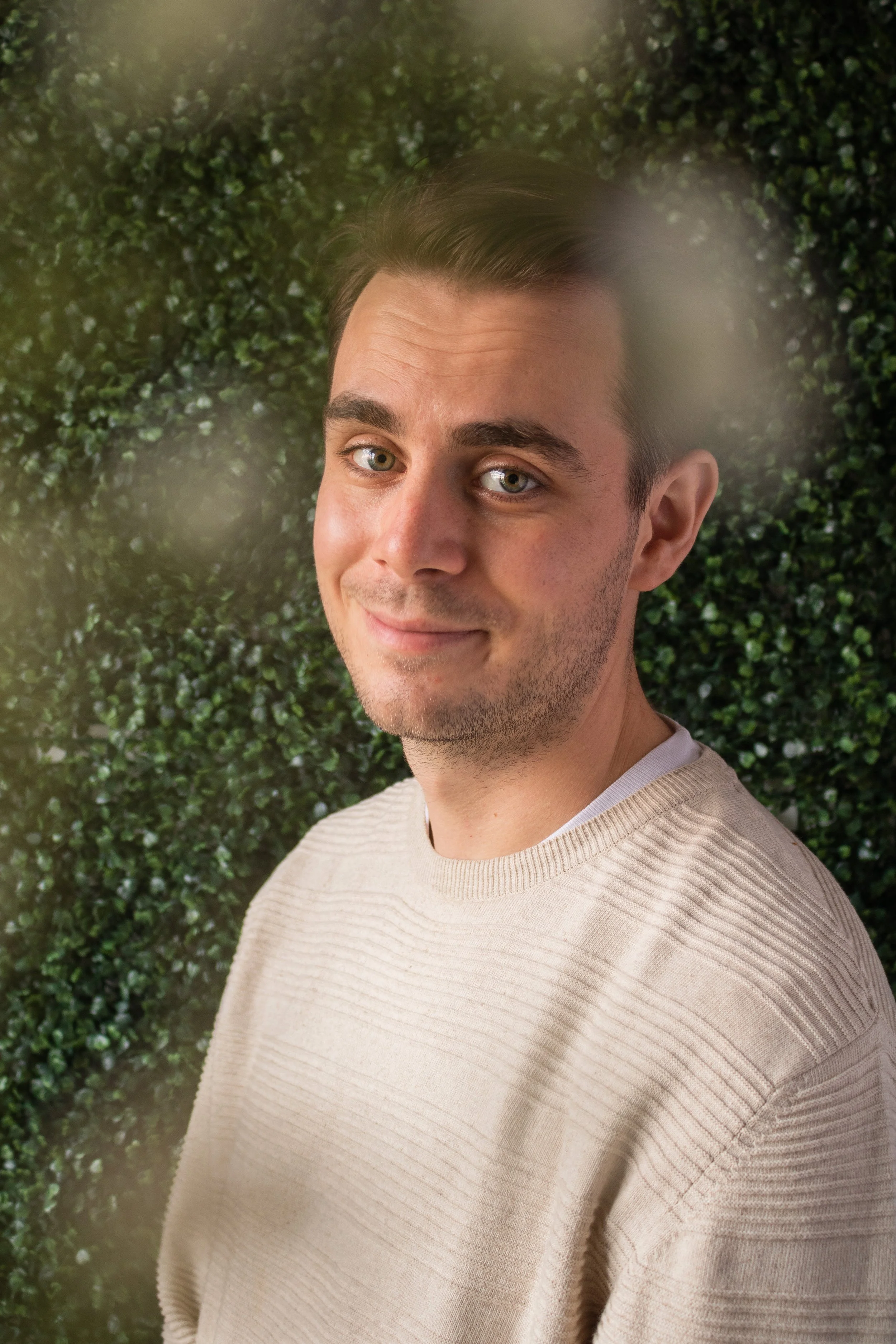 Jack, a white man, smiles at the camera. He has short brown hair, and is wearing a cream sweater. Around the edge of frame are small blurry white flowers, the background is green leaves.
