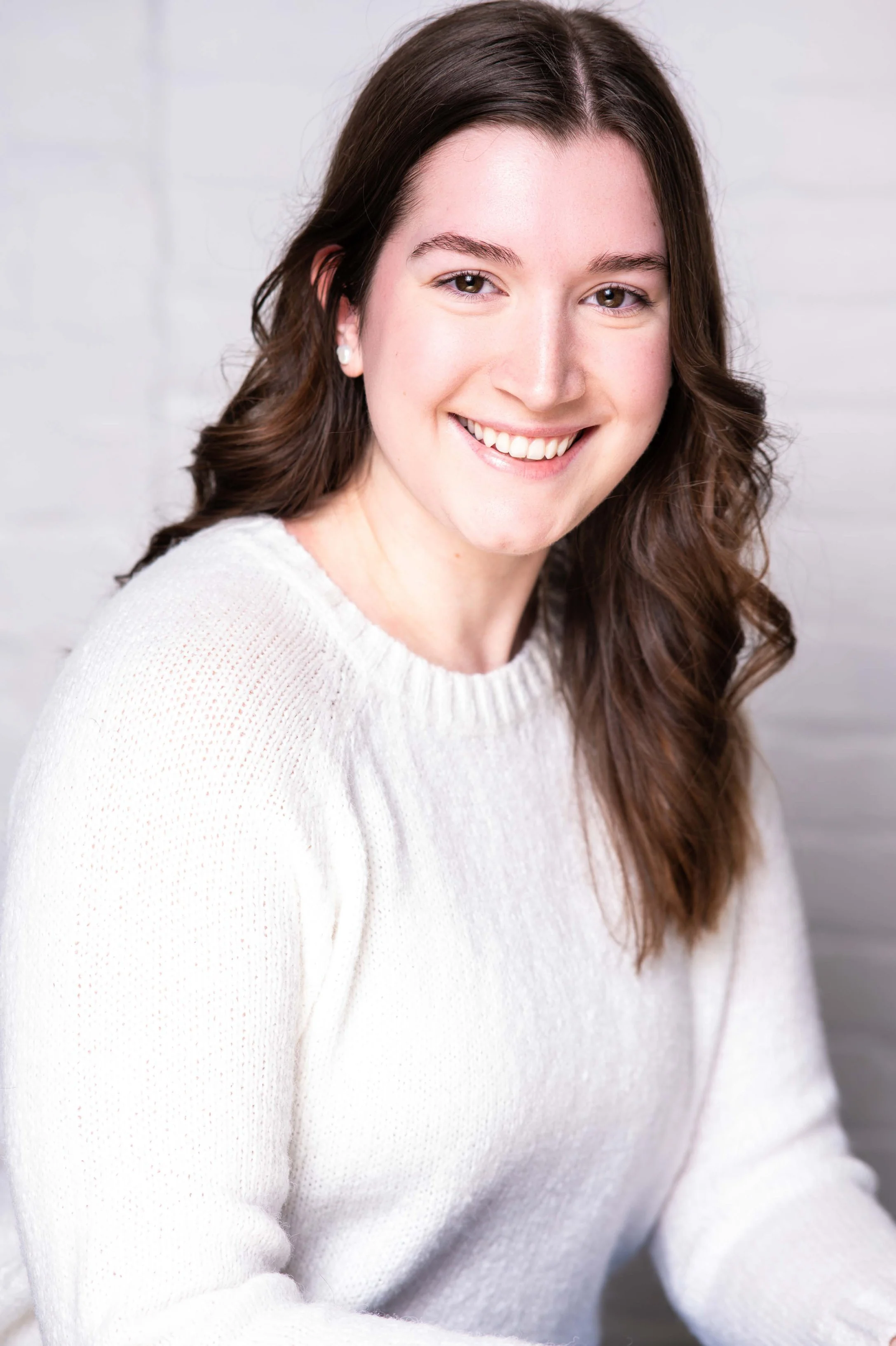 Caillen is a white woman in front of a white brick background. She has long brown hair, and is smiling at the camera. She is wearing a white sweater.