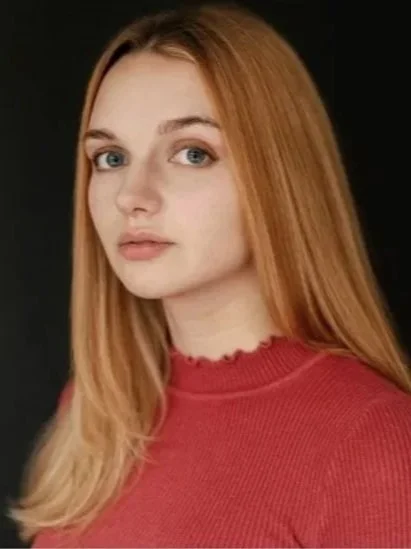Madi, a white woman with blue eyes and long blonde hair looks to camera. She is wearing a coral shirt and is in front of a black background