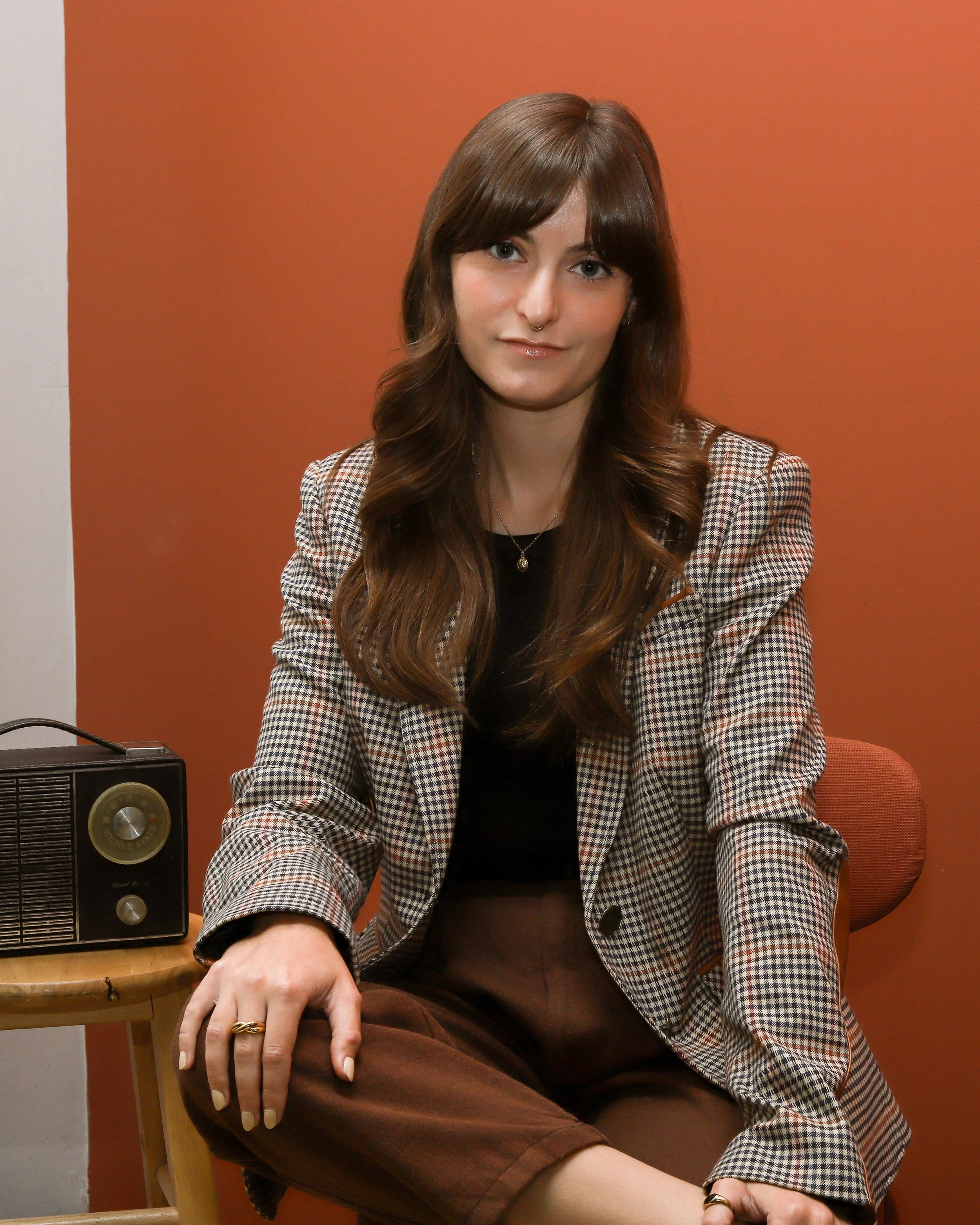 Riley, a white person, is sitting facing the camera. They wear a plaid blazer, black shirt, and brown pants. To her left, an old radio sits on a stool. The background is terracotta.