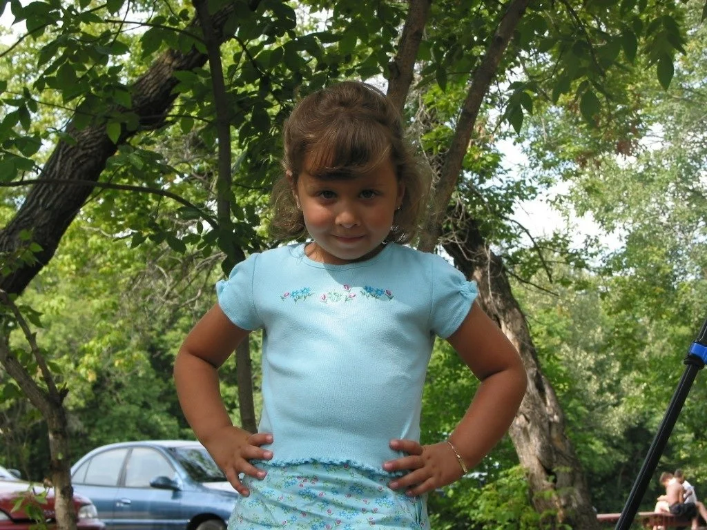 Little Josephine is standing with her hands on her hips, looking determinedly at the camera. She has short brown hair and bangs, and is wearing a blue tank top. The background is green trees.