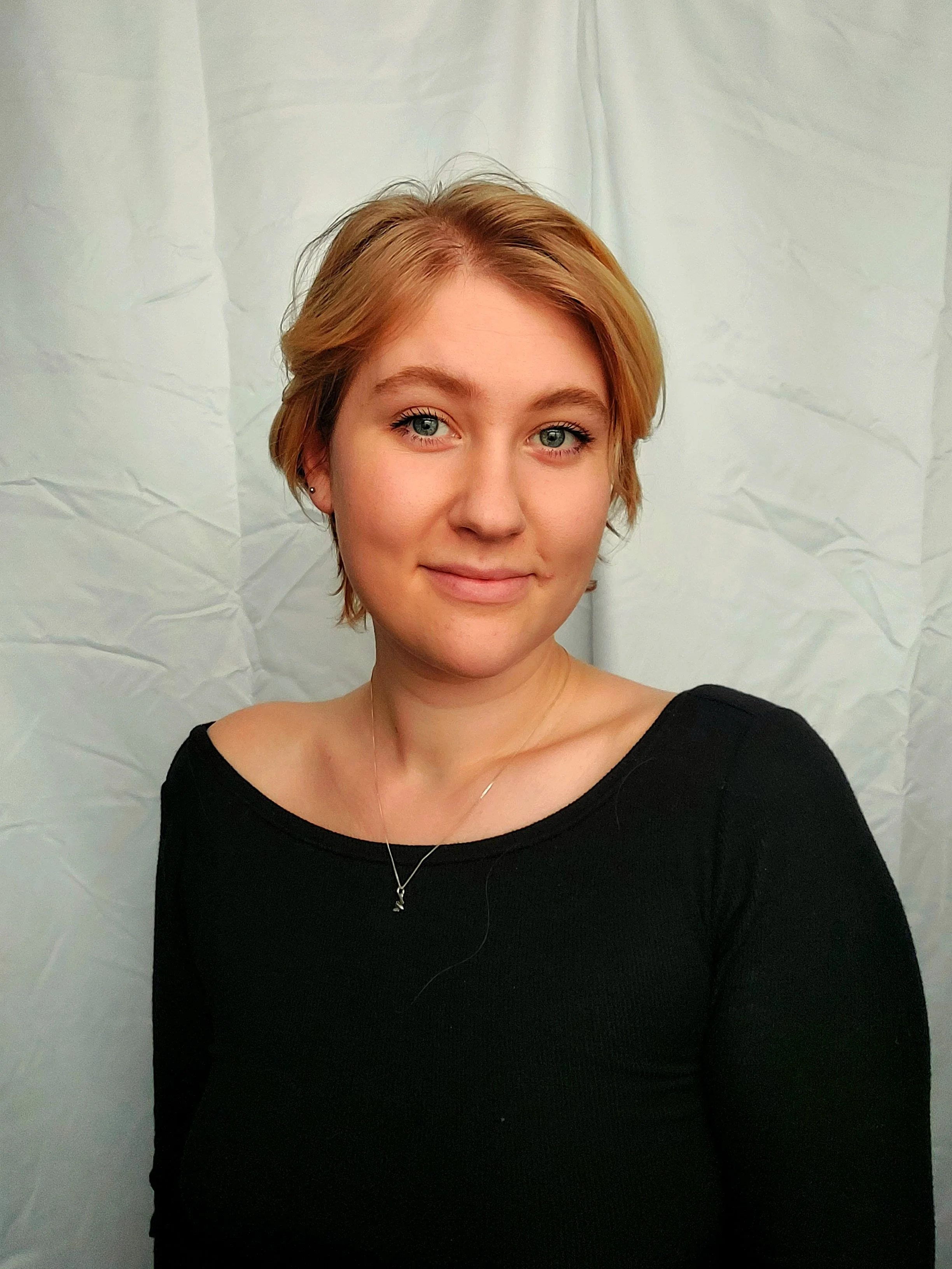 Hannah, a white woman, has short blonde hair and blue eyes. She is smiling at the camera with a white background. She is wearing a black sweater