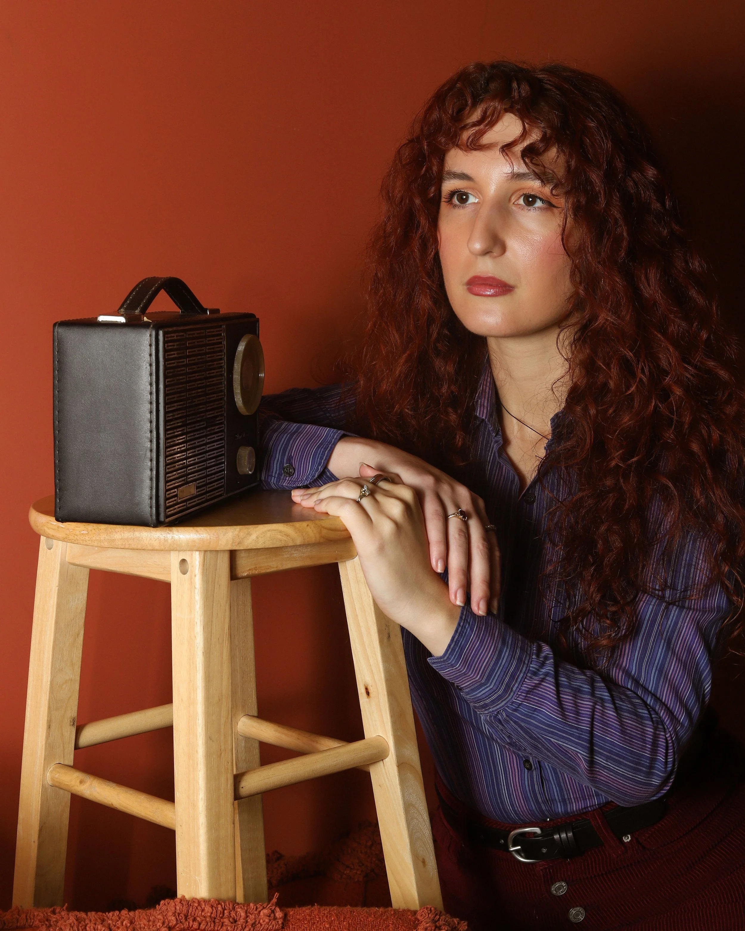 Kira, a white woman with long red hair looks off to the left of frame with a determined expression. She is resting her hands on a stool with an old radio. She is dressed in 1960s clothing, against a terracotta background