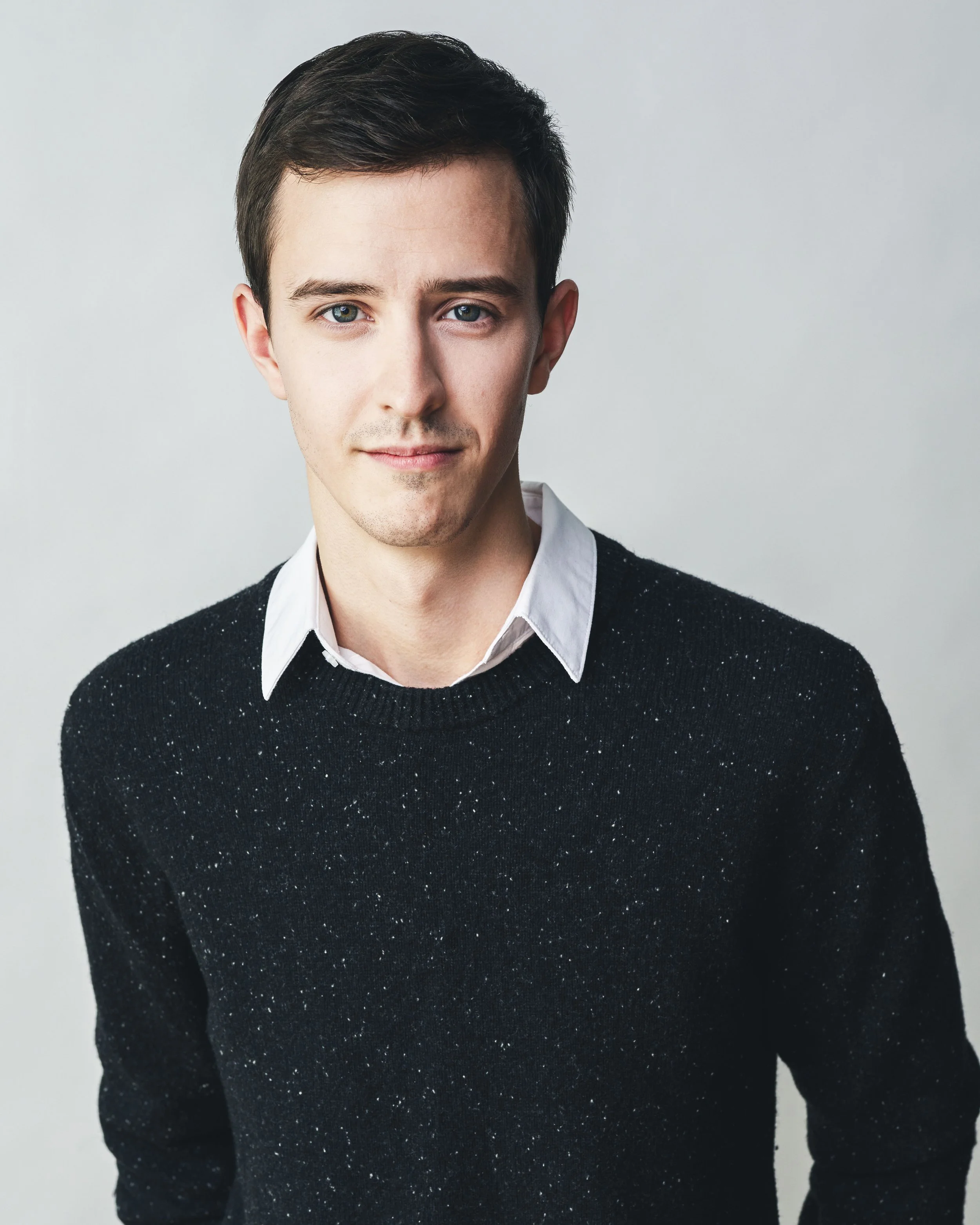 Adrian, a white man, stands in front of a white background. He has short brown hair and blue eyes. He is wearing a navy sweater, and the collar of a white shirt sticks out the neck of the sweater.