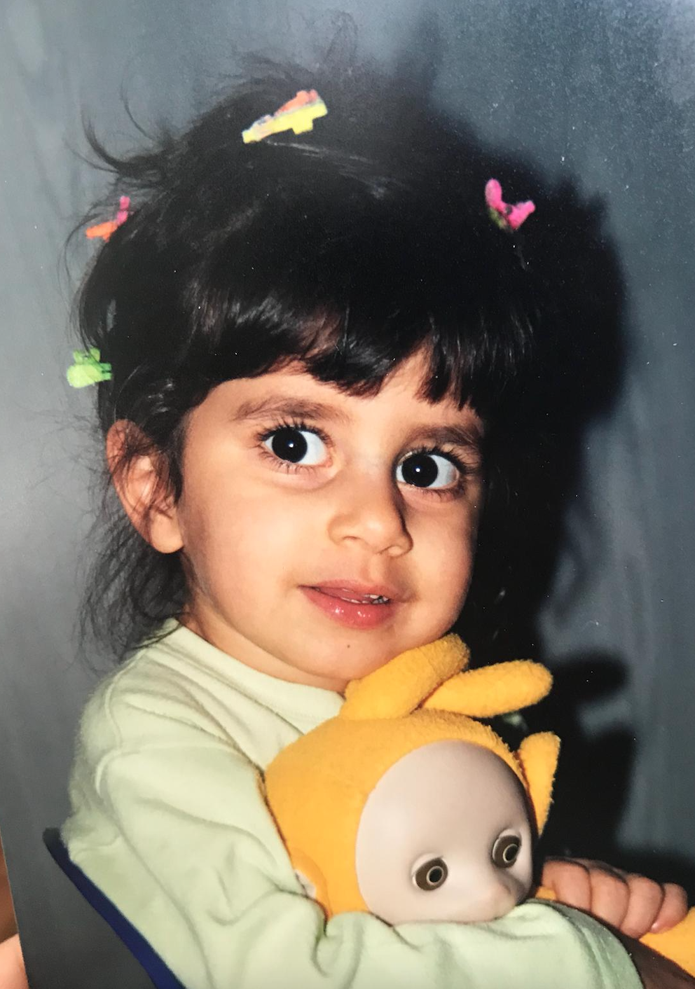 Little Elif is looking warily at the camera, wearing multi-coloured clips in her hair and a green shirt. She is holding a yellow teletubby doll.