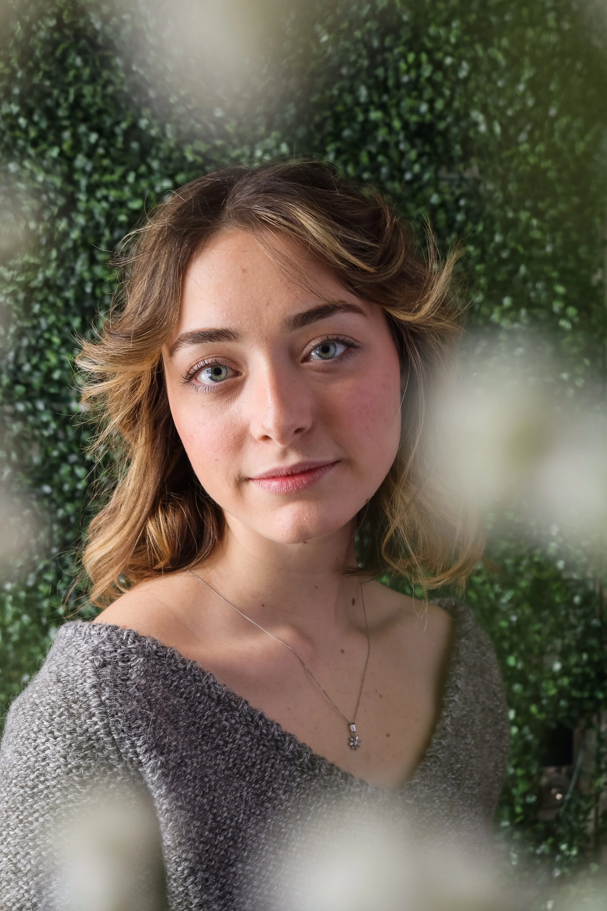 Lucy, a white woman, is smiling softly at the camera. She has short blonde hair and blue eyes. She is wearing a grey sweater. Around the edge of frame are small white blurry flowers, the background is green leaves.