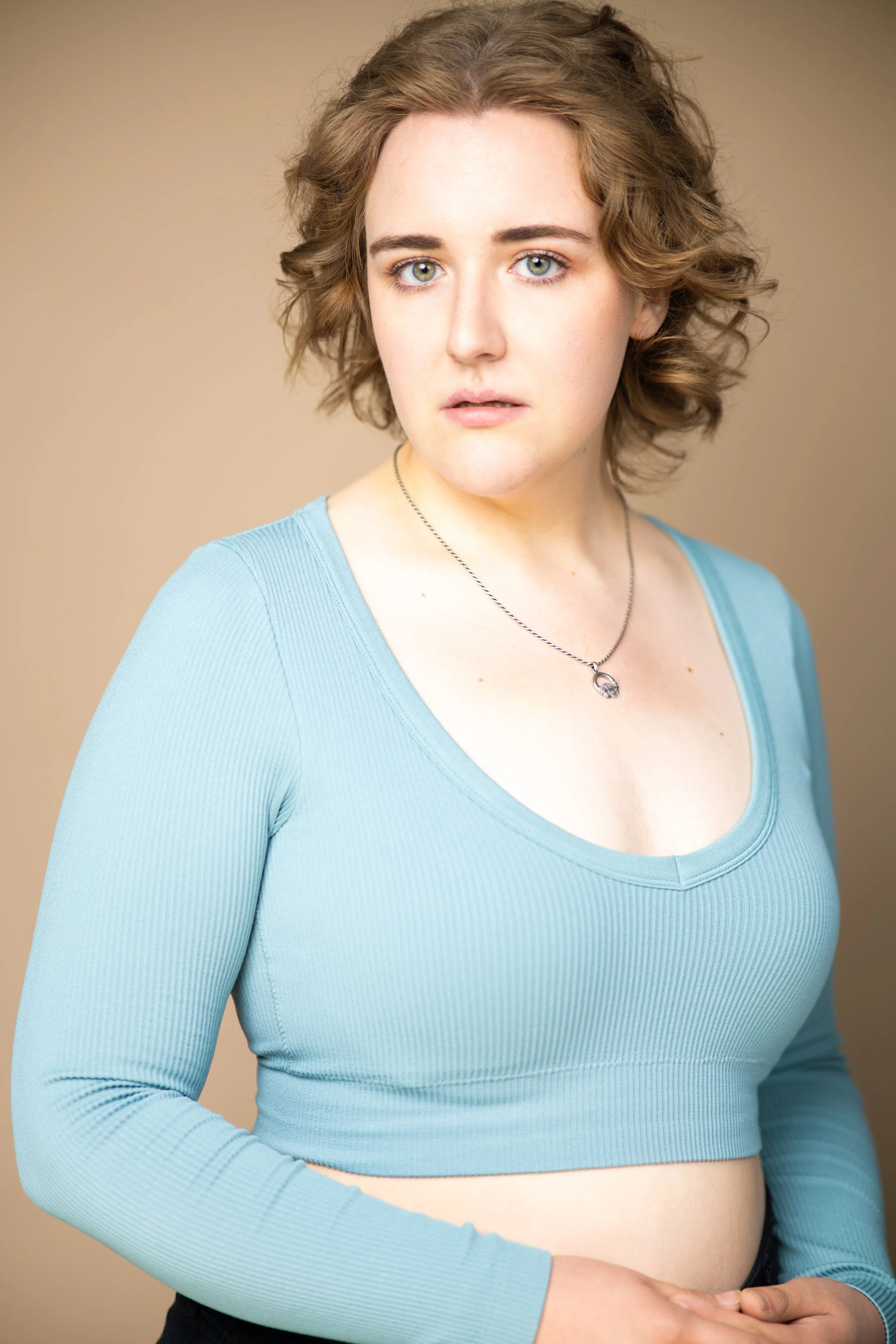 Bronwyn, is a non-binary female presenting performer standing in front of a light brown background. They. have short brown. hair and blue eyes, and are looking to camera. She is wearing a blue long sleeved crop top and black pants.