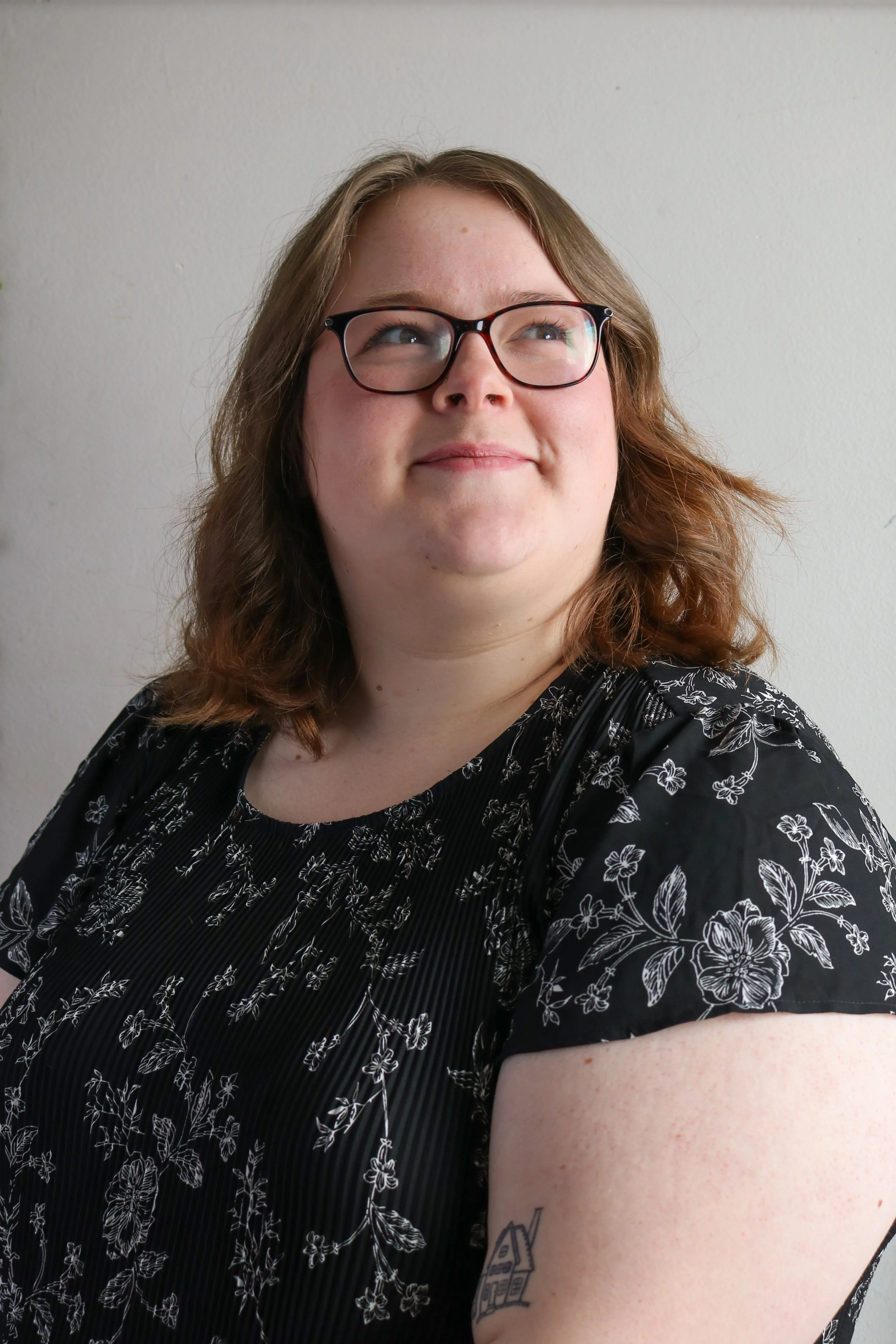 Emily, a white person, is smiling at something out of frame. She has shoulder length brown hair and glasses. They are wearing a black floral blouse. The background is white.