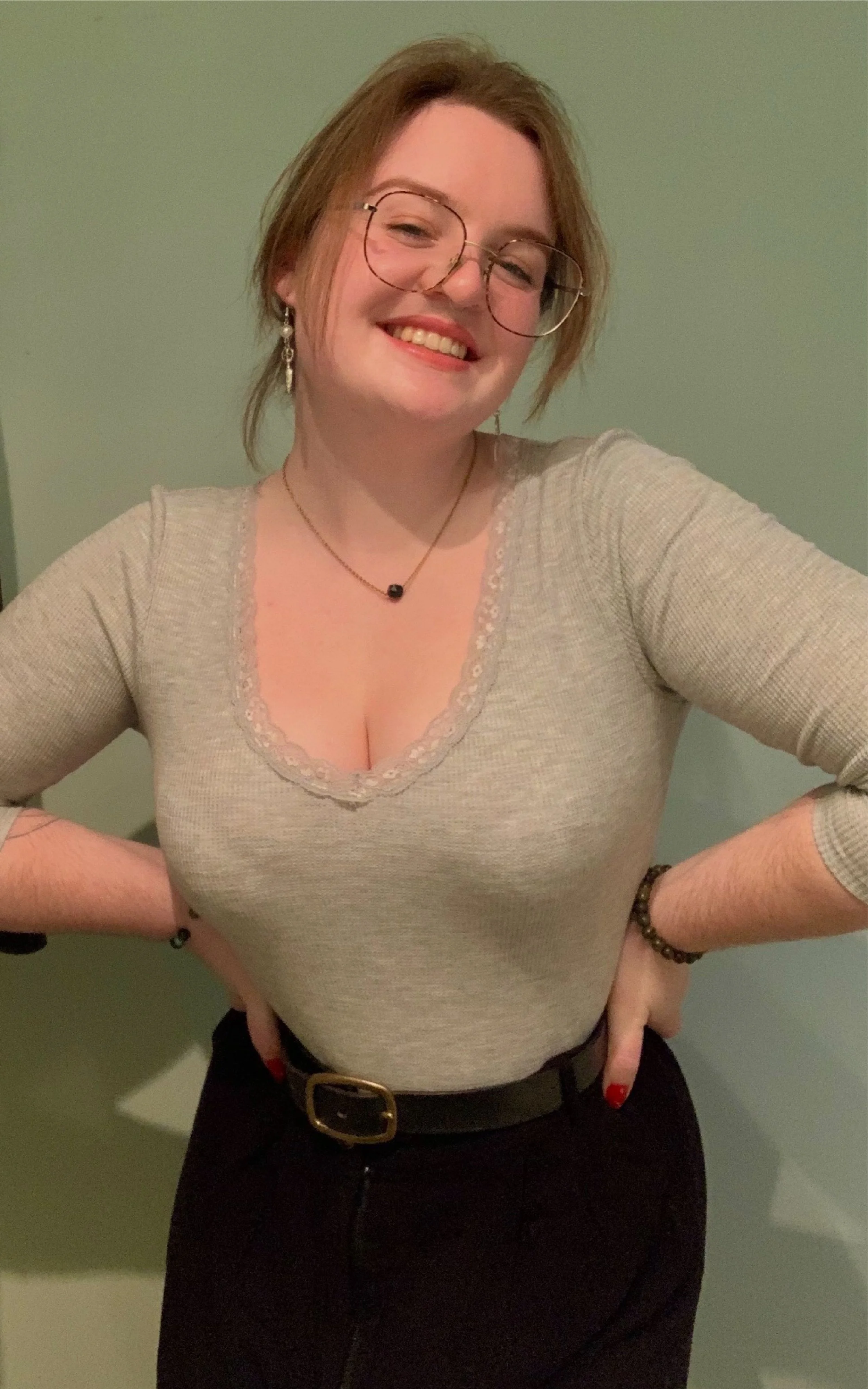 Julianna, a white non-binary performer is standing in front of a green wall, smiling at the camera. They are wearing glasses, a grey long sleeve shirt and black pants. Her hair is pinned up behind her head.