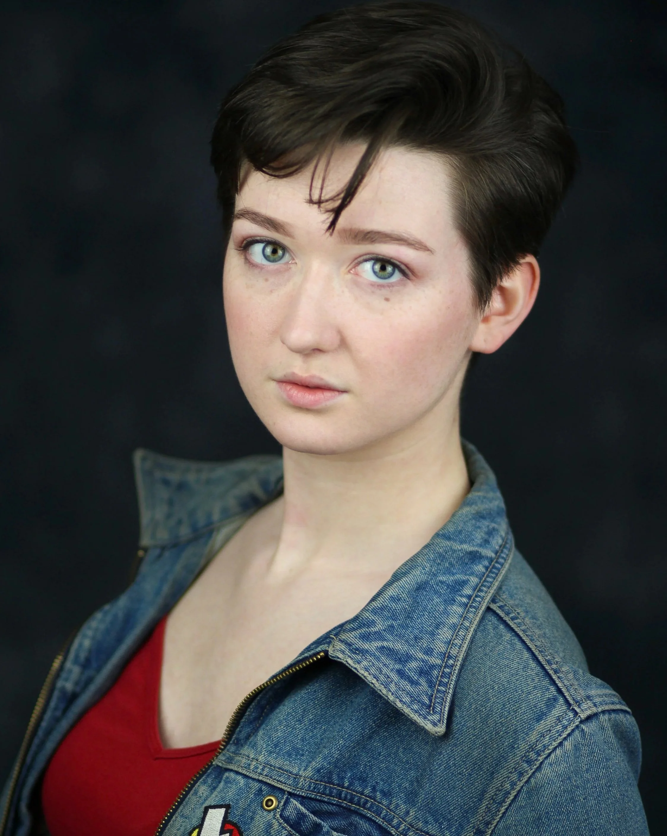 Alison, a white non-binary performer, is in front of a black background. They have short brown hair and light blue eyes. She is wearing a red shirt and denim jacket.