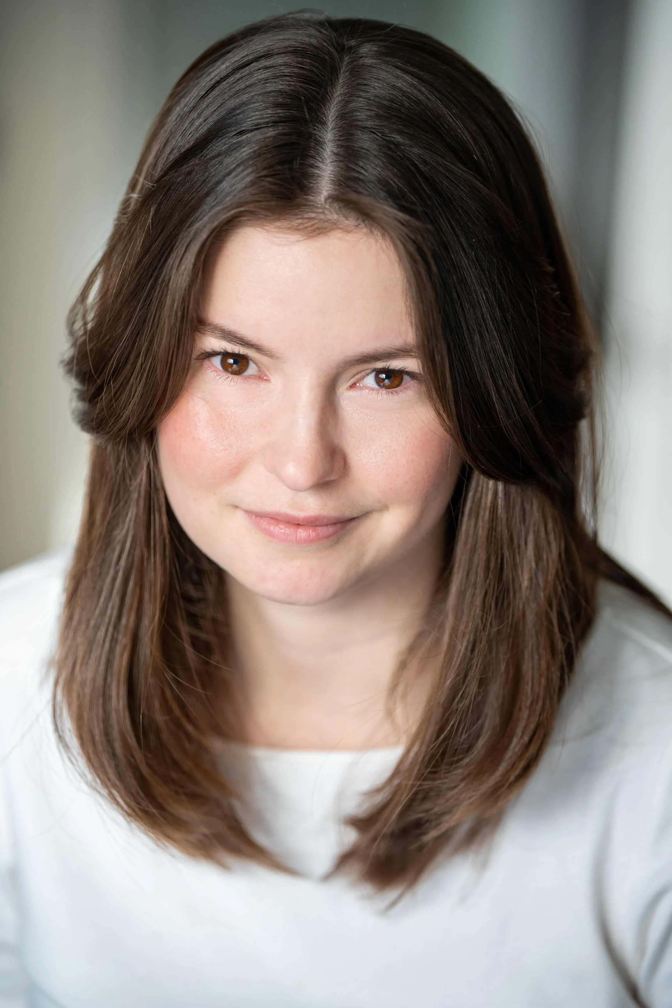 Evelyne, a white woman, smiles softly at the camera. She has shoulder-length brown hair and is wearing a white blouse. The background is light grey.