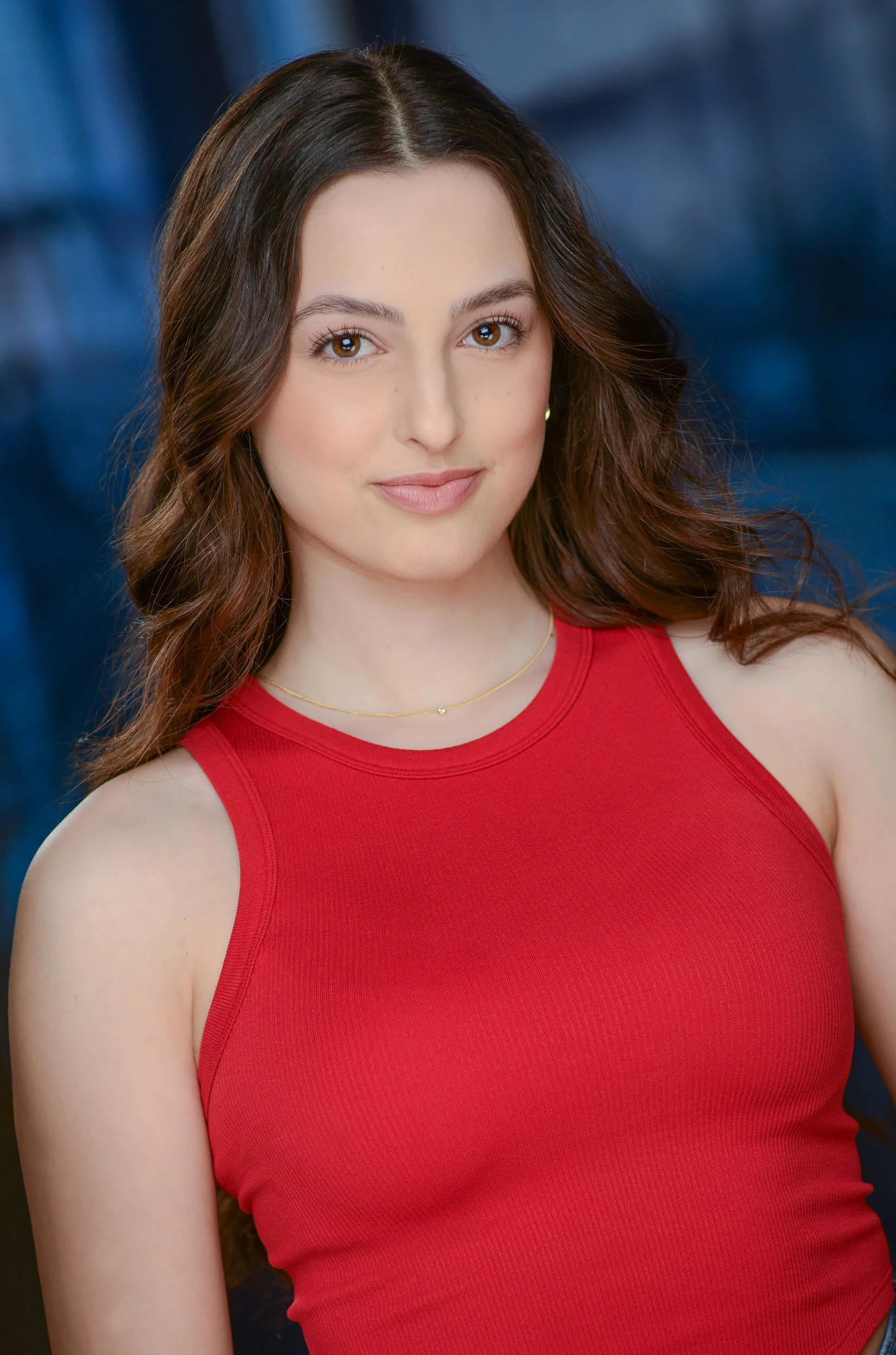 Chloe, a white woman, is smiling slyly at the camera. She has long brown hair and is wearing a red tank top. The background is blue.