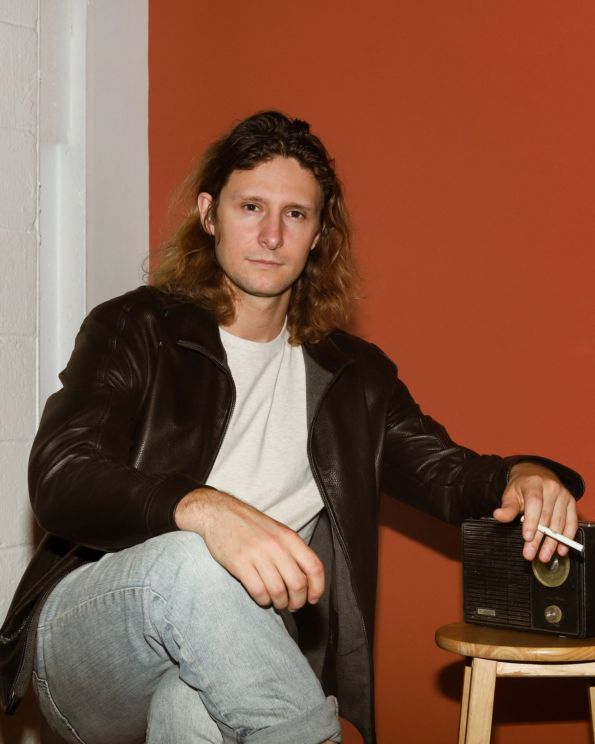 Erik, a white man, is seated, wearing a leather jacked, white shirt, and blue jeans. He is leaning his arm on a stool beside him. The background is terracotta.
