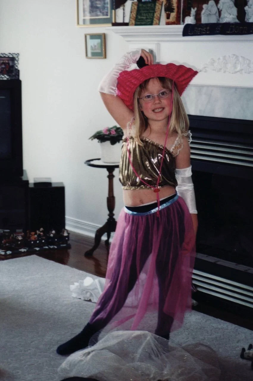 Little Rachel is smiling at the camera, posing in front of the fireplace. SHe has on a red cowboy hat, long opera gloves, a shiny gold tank top, and a long purple tutu over black tights.