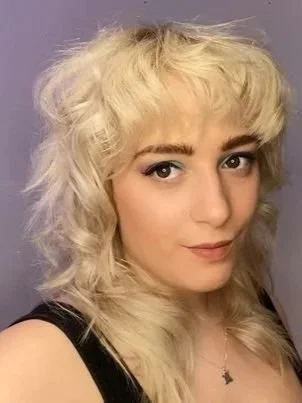 Sabrina, a white woman, smirks at the camera. She has blonde shaggy hair and brown eyes. She is wearing a black top, and stands against a purple background