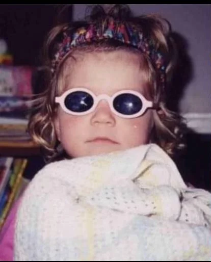 Little Mari is looking a the camera with a neutral expression. She is wearing a tiara as a headband, and round sunglasses. She is holding a knitted baby blanket.