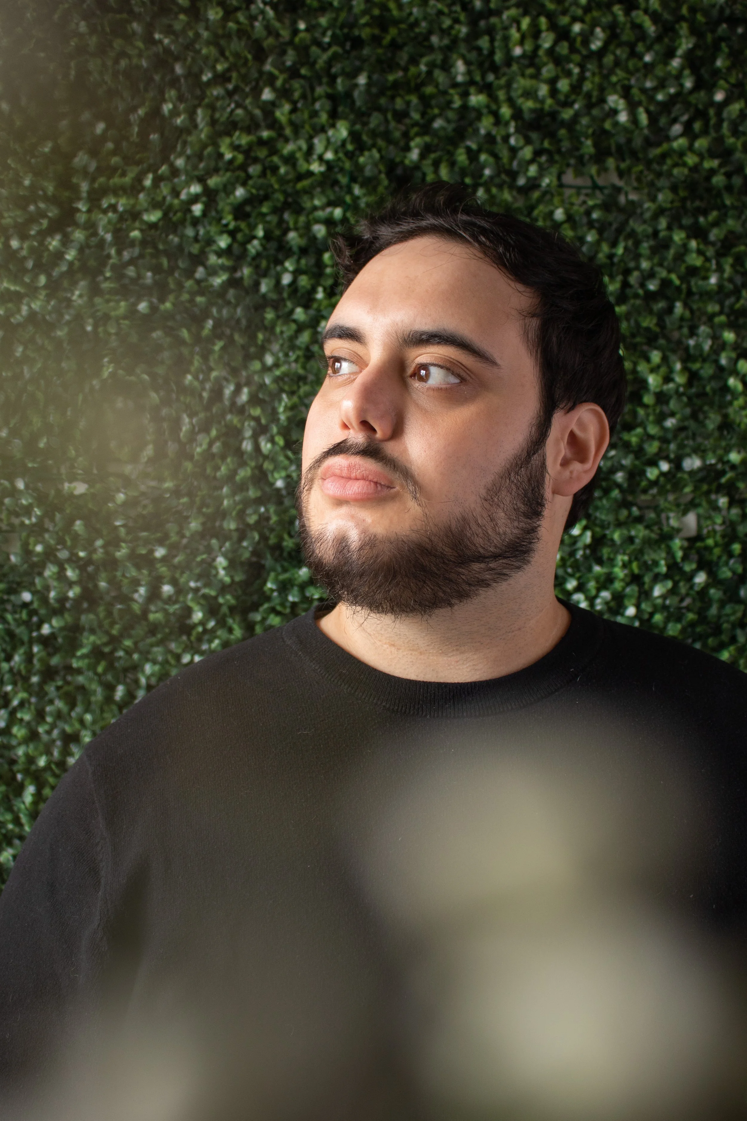Matt is looking off into the distance. He has short black hair and a beard. He is wearing a black t-shirt. Around the edge of frame are small blurry white flowers, the background is green leaves