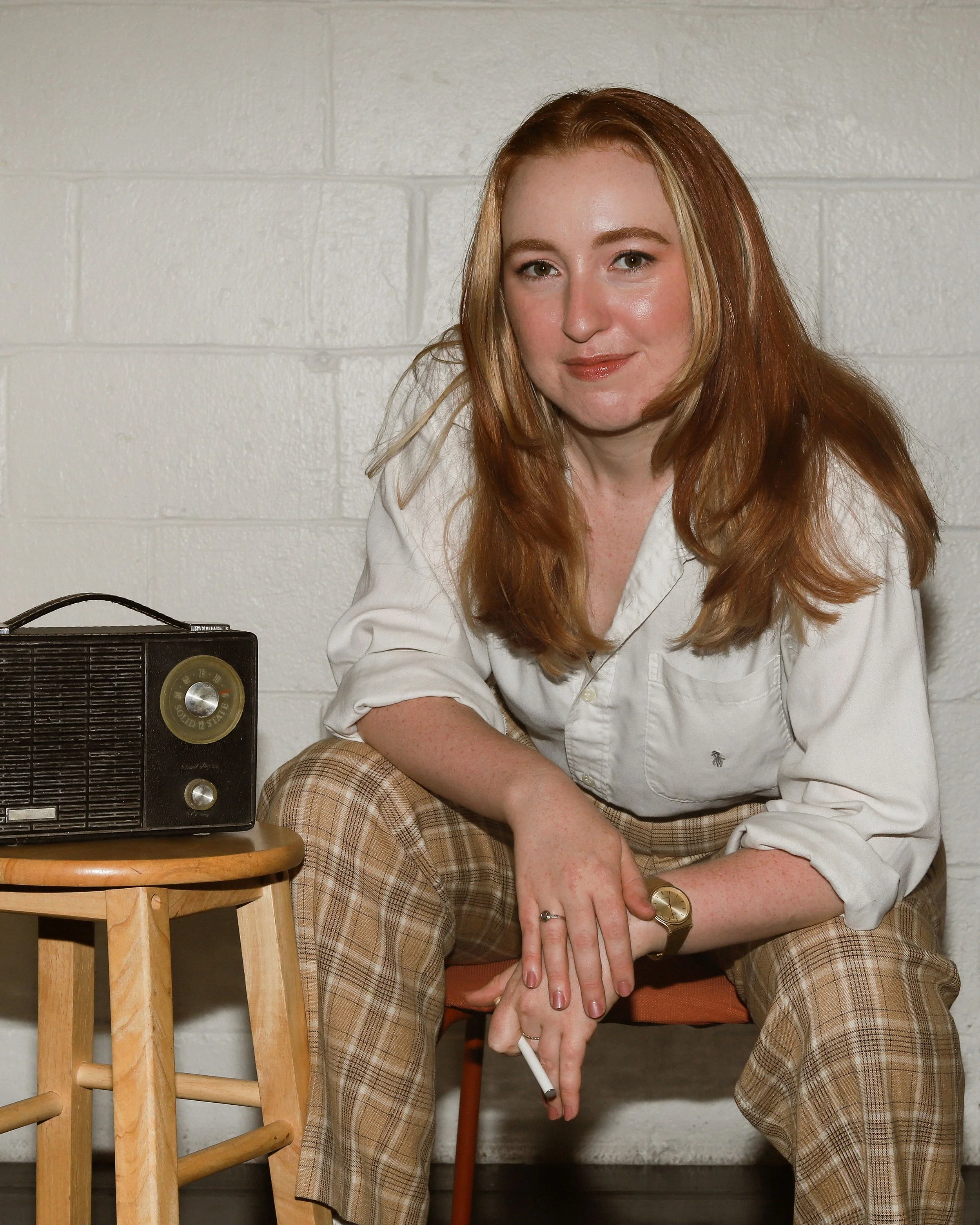 Katherine, a white woman with long red hair, leans towards the camera conspiratorially. She is wearing plaid pants, a white button down, and has a cigarette in her hand. To her left, an old radio sits on a stool. The background is white brick.