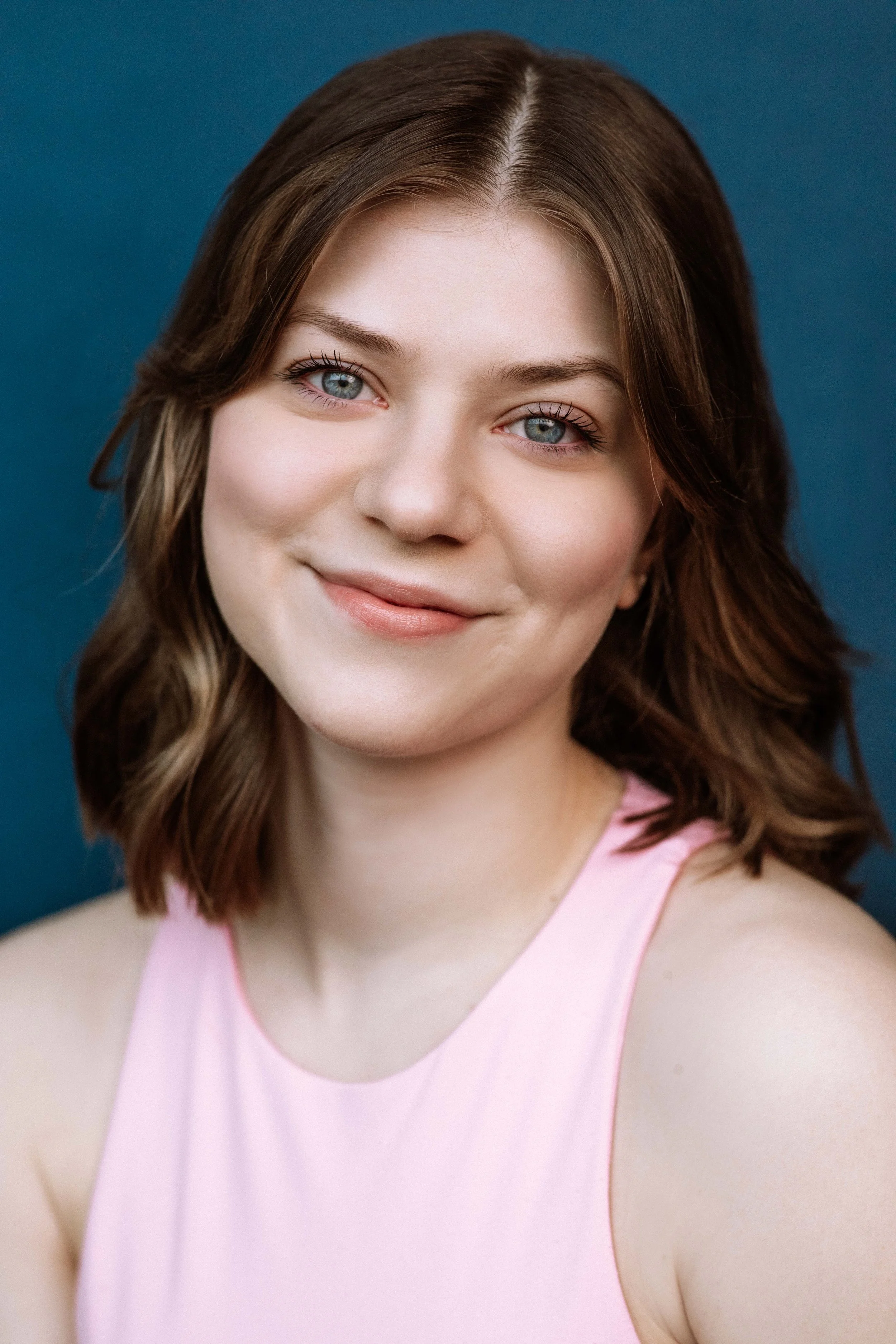 Mari, a white woman, is smiling softly at the camera. She has shoulder-length brown hair and blue eyes, and is wearing a pink tank top. The background is teal.