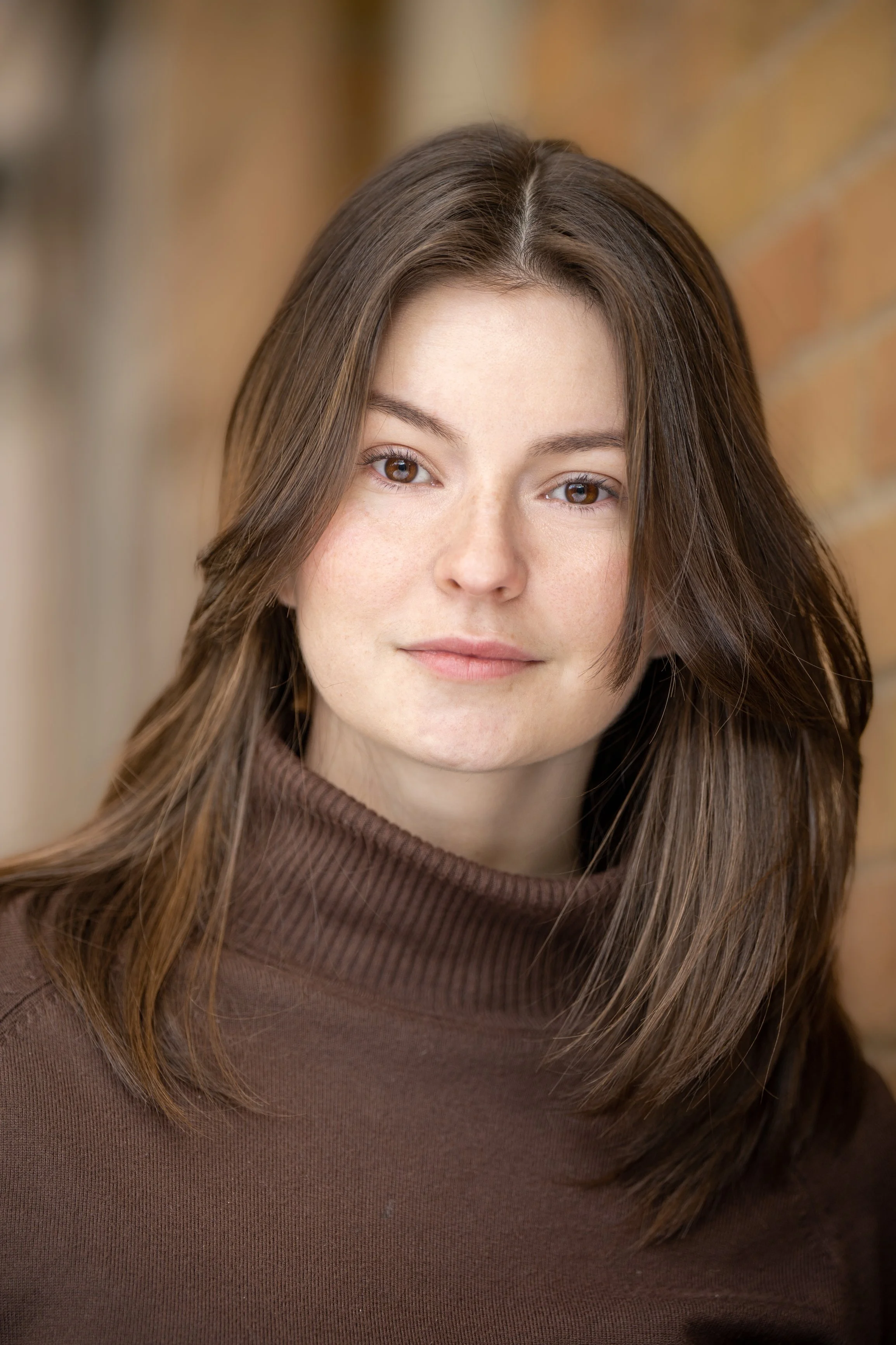 Evelyne, a white woman, looks to camera with a neutral expression. She has shoulder-length brown hair, and is wearing a brown turtleneck. The background is red brick.