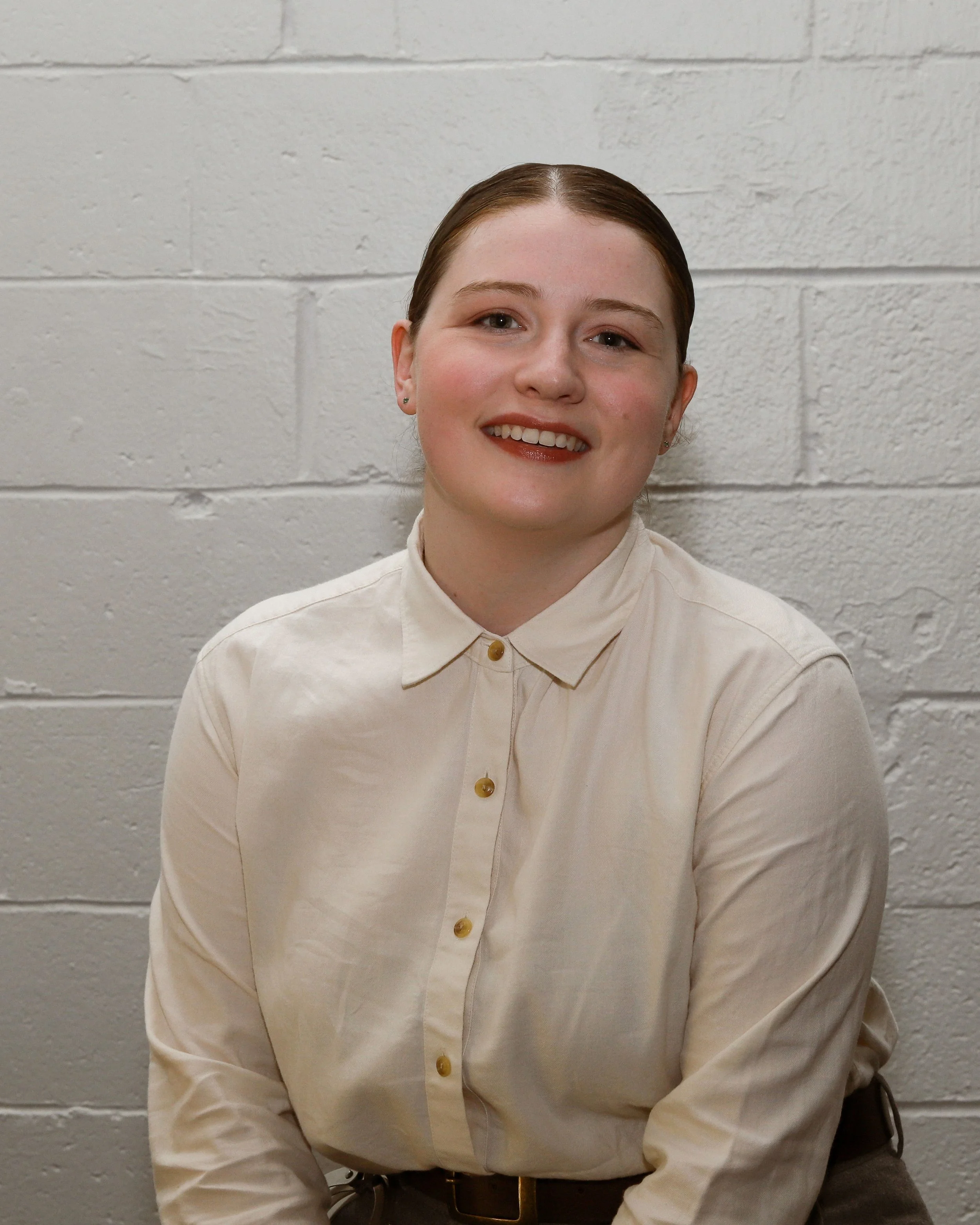 Fiona, a white woman, smiles at the camera. Her red-brown hair is pulled tightly against her head to a bun. She is wearing an off-white button down shirt. The background is white brick.