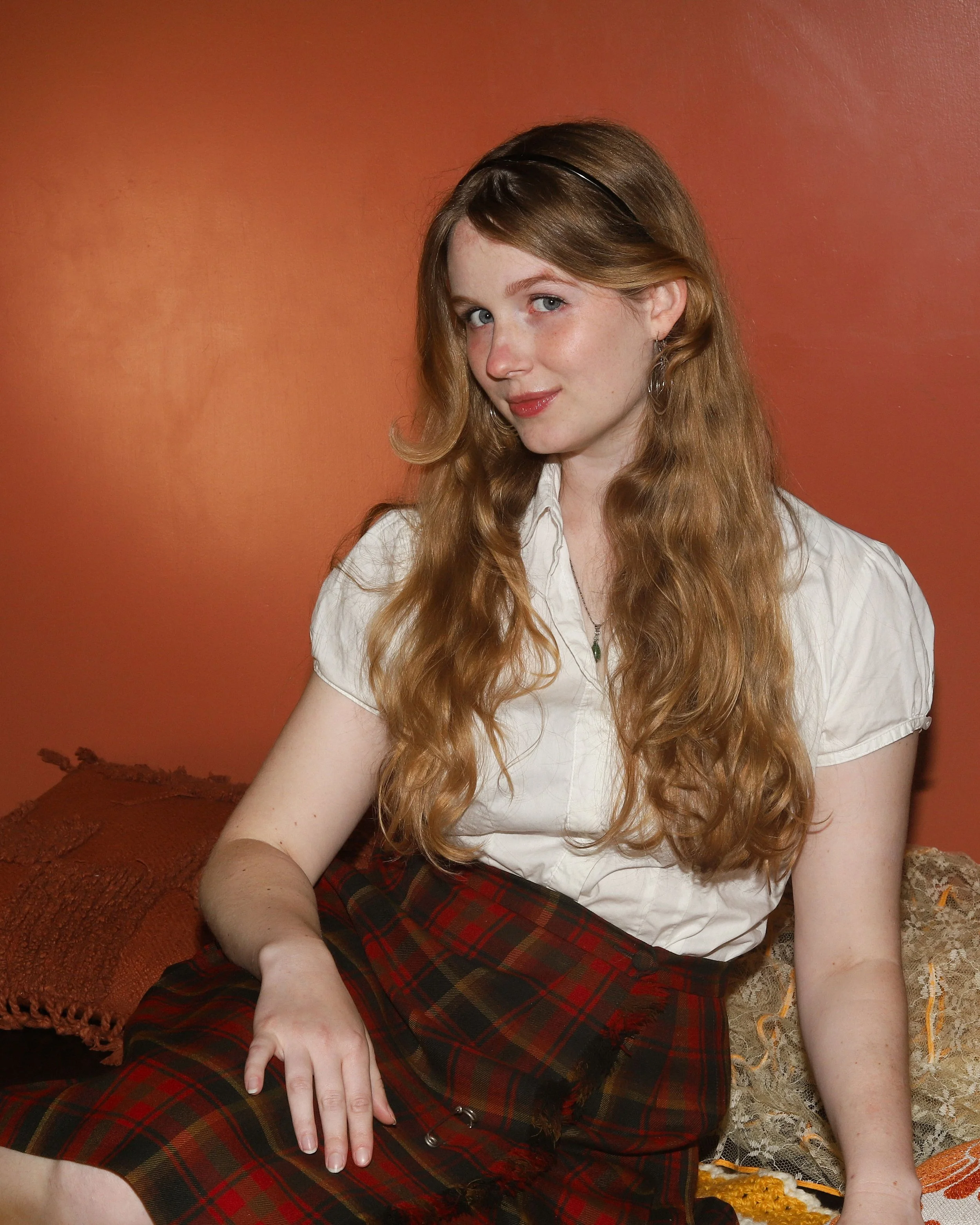 Columbia, a white woman with long blonde hair, looks with a sly smile at the camera. She is seated, and wearing a plaid skirt and white top against a terracotta backdrop.