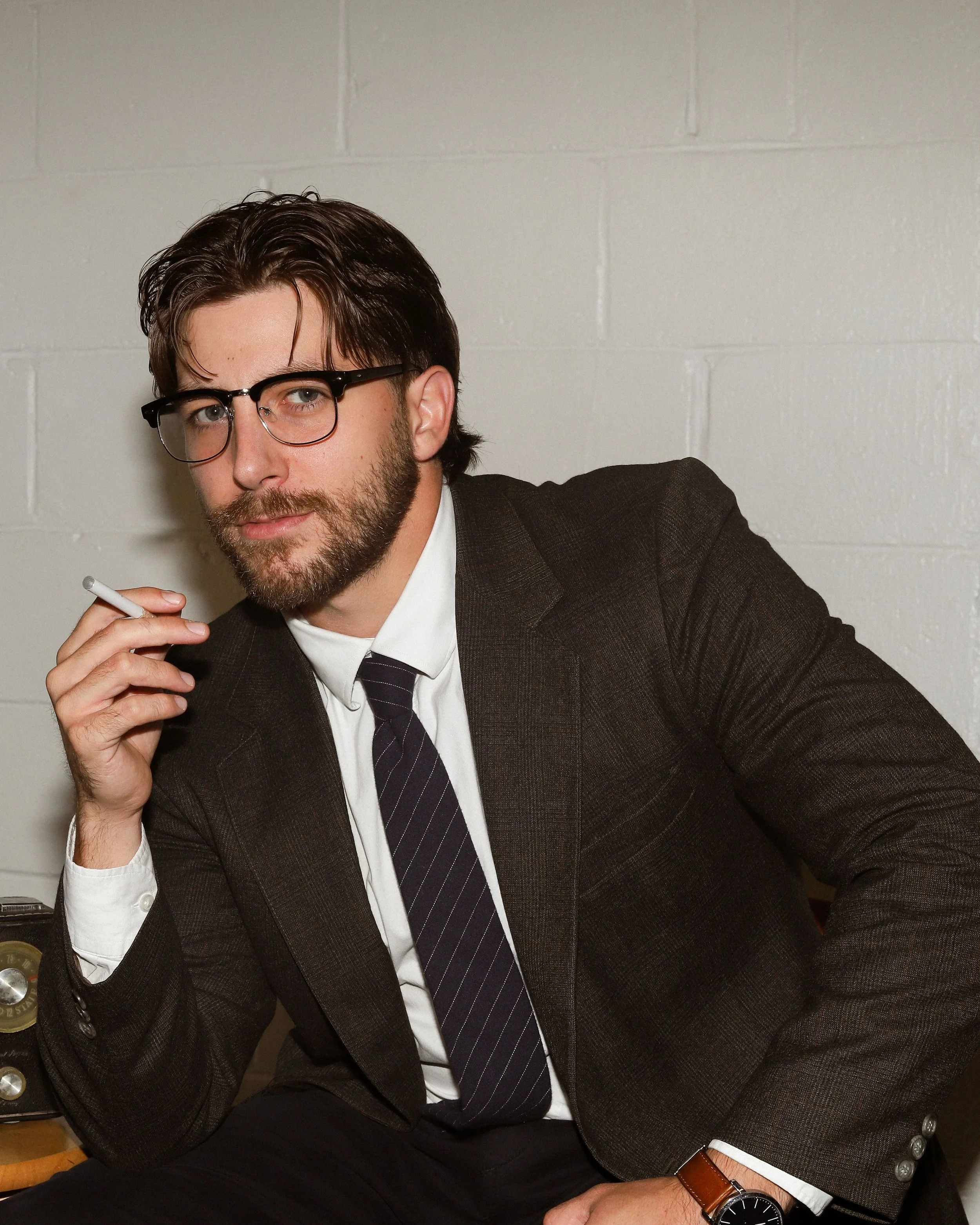 Lucas, a white man, leans towards the left of frame with a cigarette in his hand. He has brown hair and a beard. He wears glasses and a charcoal suit with a blue tie. . The background is white brick.