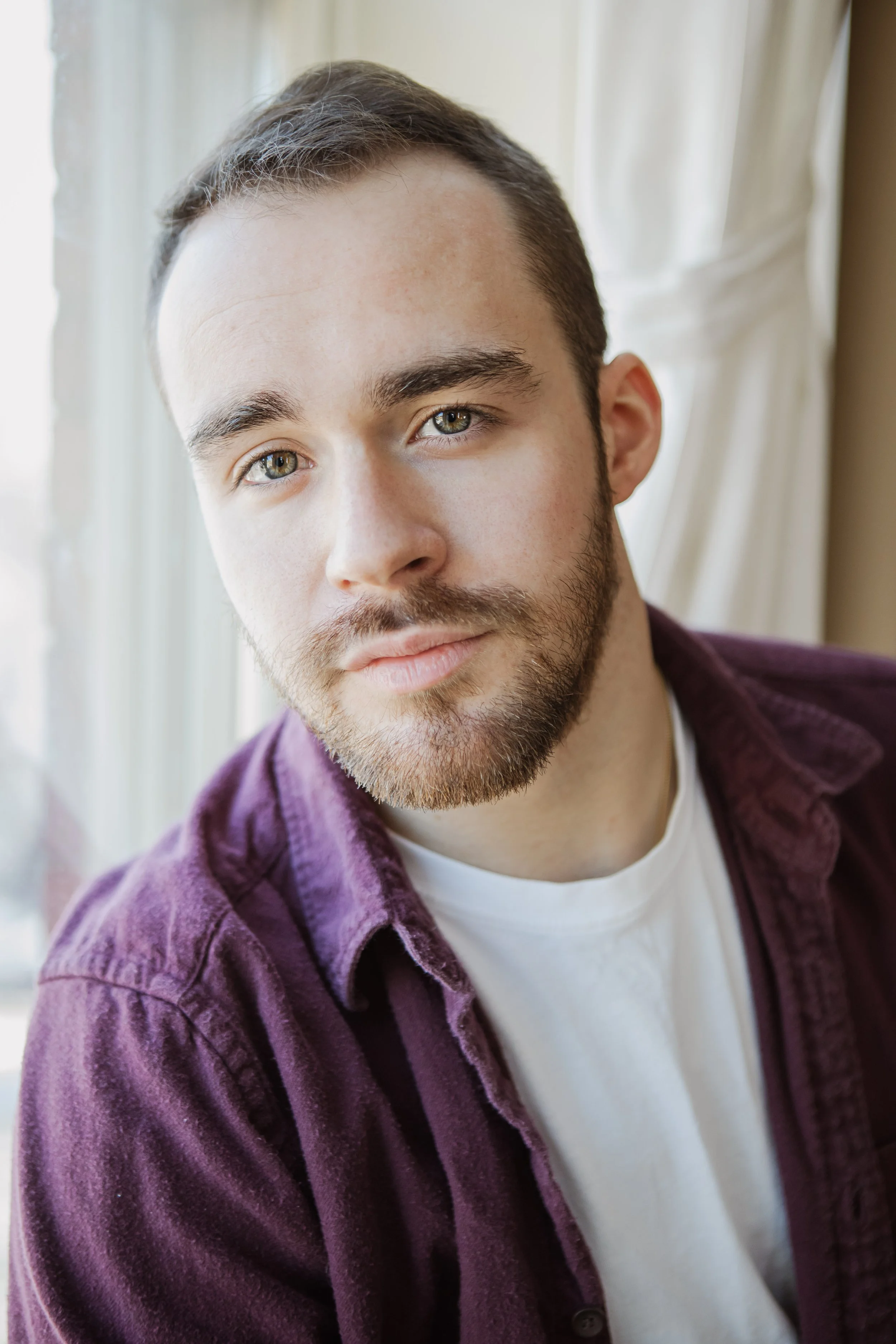 Joseph, a white man, leans slightly to the left but is looking at the camera with a pleasant neutral expression. He has close-cropped brown hair and a beard, and is wearing a purple button down over a white t-shirt