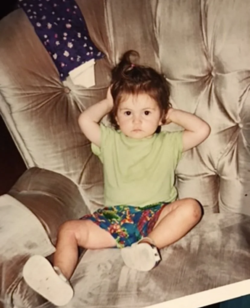 Little Evelyne sits in a tufted brown chair, with her hands over her ears. She has short brown hair, and is wearing a green t-shirt, multicoloured shorts, and running shoes.