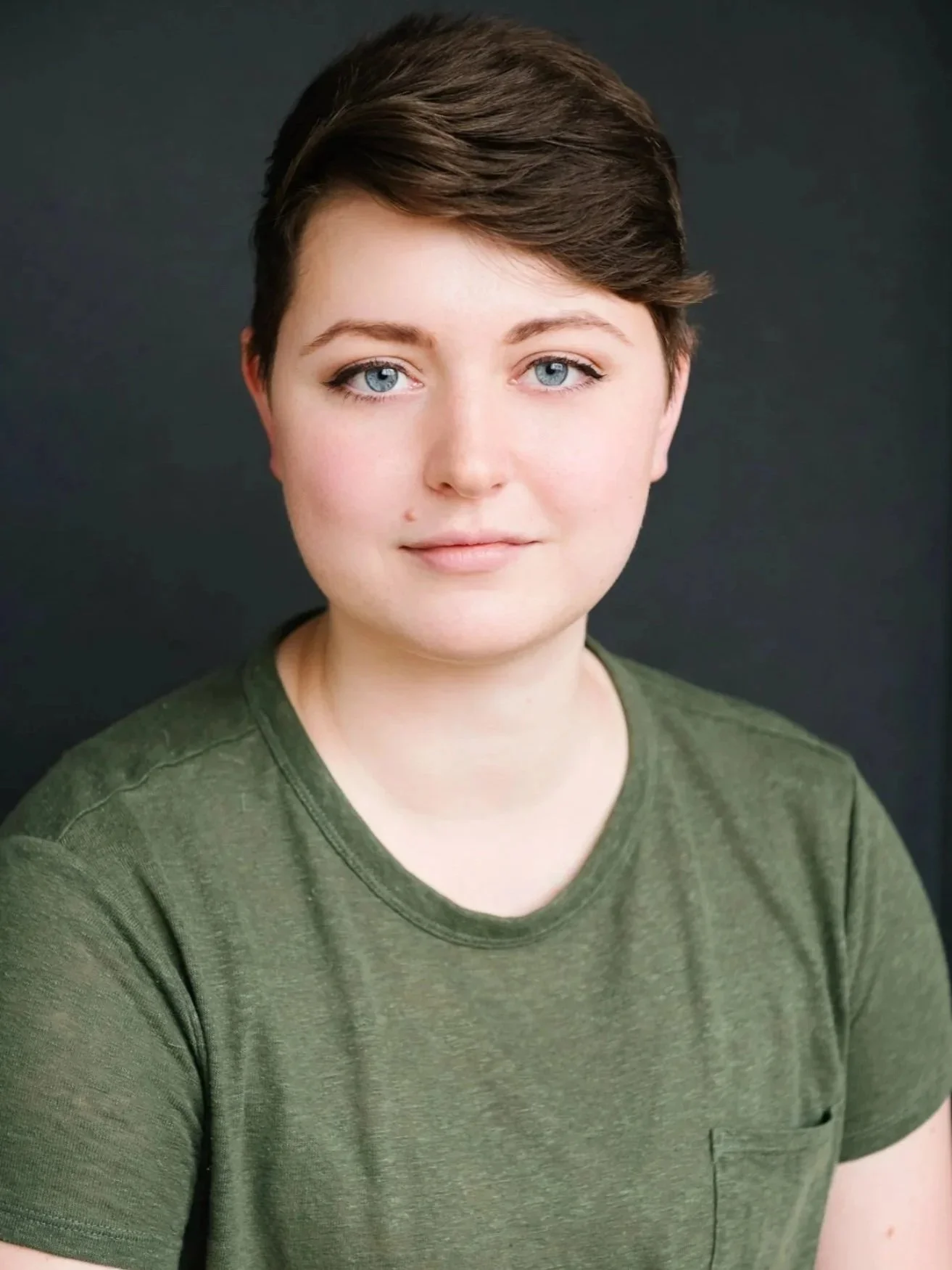 Hadley, a non-binary white performer, is in front of a grey background. They have short brown hair and green eyes, that match their green t-shirt.