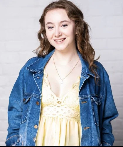 Sydney, a white woman, is smiling at the camera. She has long brown hair, and is wearing a yellow dress and a blue jean jacket. The background is white.