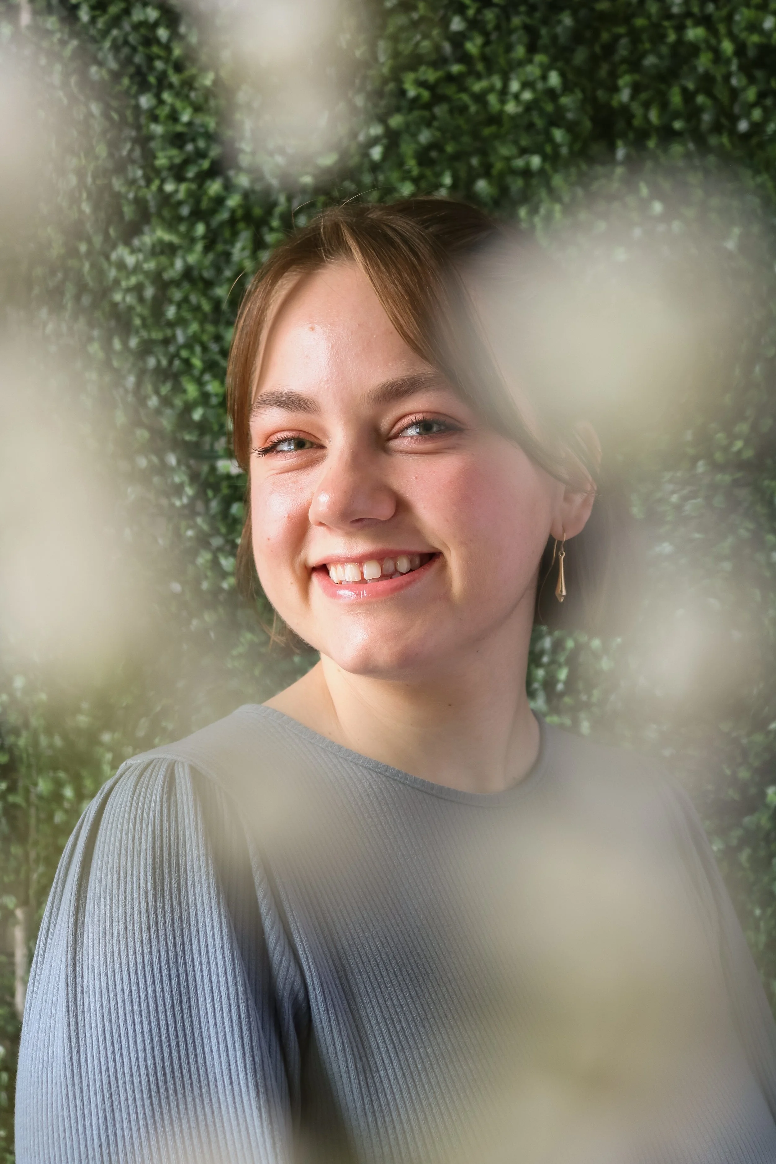 Alex, a white person, is smiling at the camera. They have a brown bob, and are wearing gold earrings. She is wearing a blue dress. Around the edge of frame are blurry white flowers, the background is green leaves.
