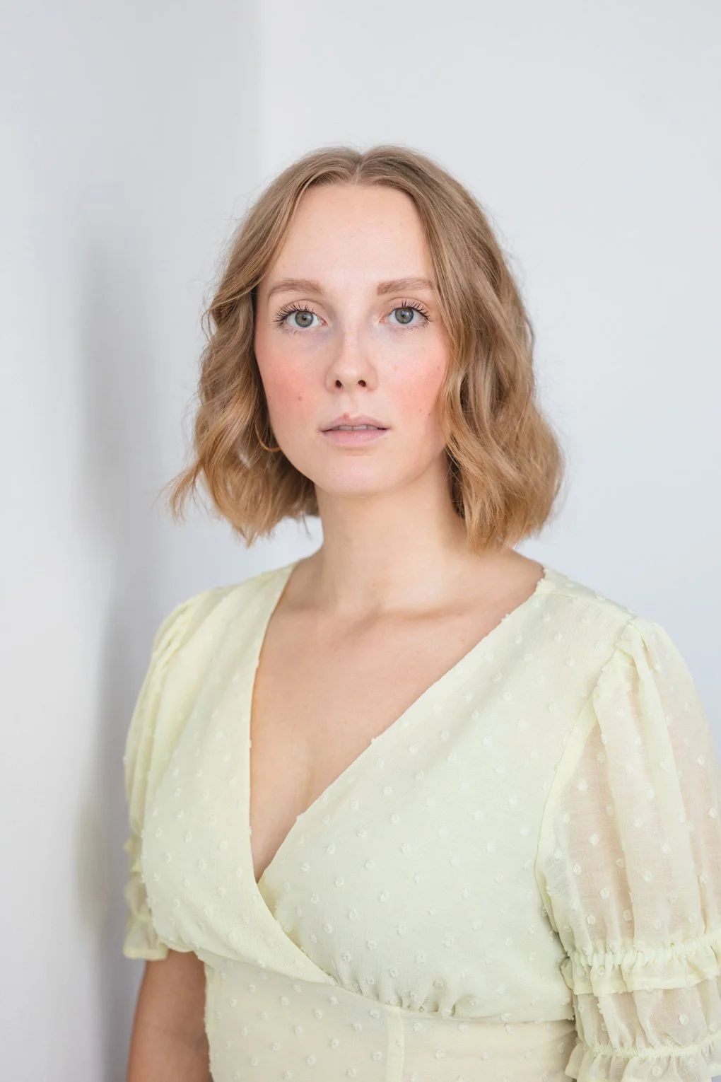 Rachel, a white woman, is looking wistfully at the camera. She has a blonde bob, and is wearing a light yellow blouse. The background is white.