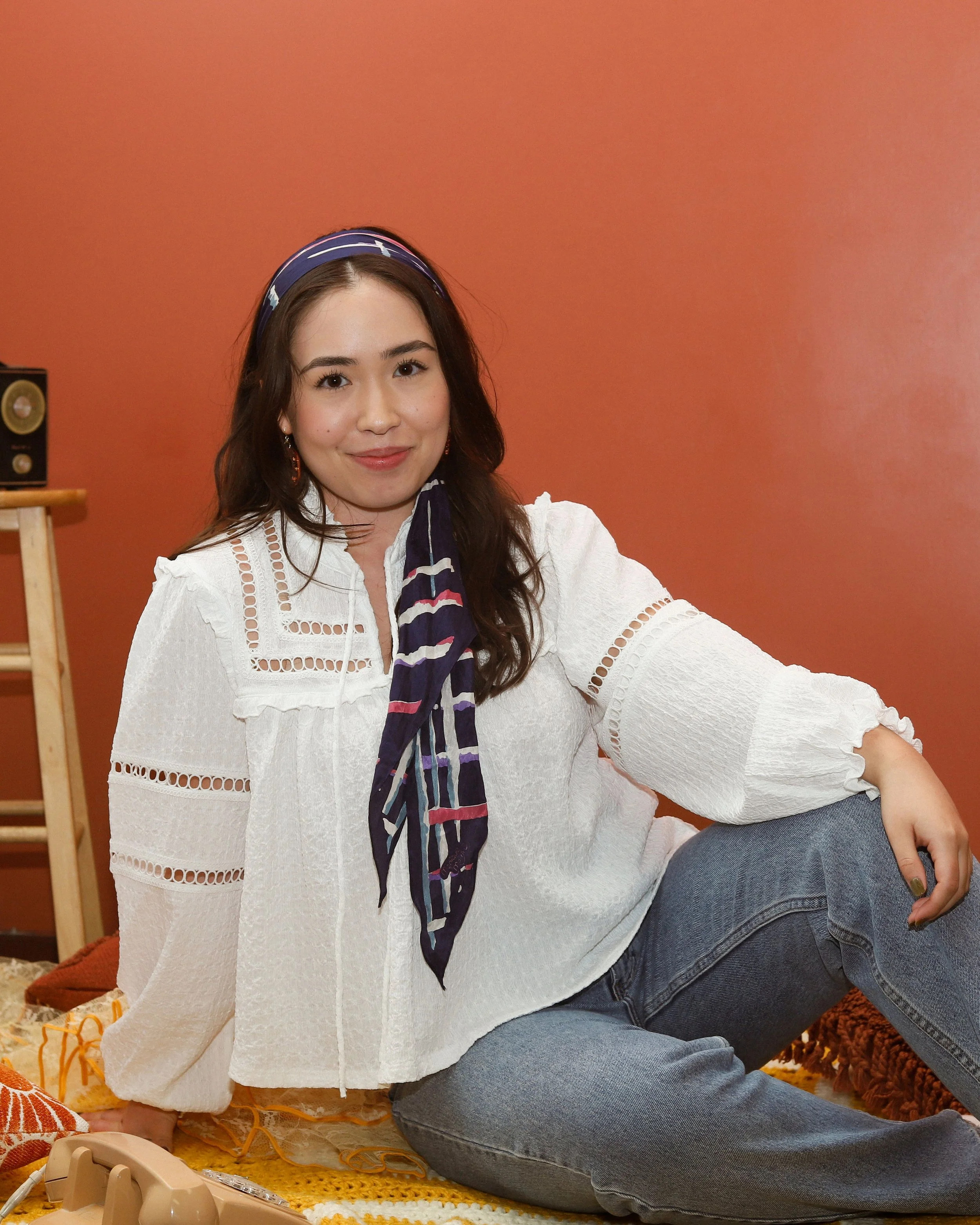 Alicia, an Aisan-Canadian woman, is seated, leaning on her arm. She is smiling at the camera. She has long black hair, and is wearing a headscarf, white shirt, and blue jeans. The background is terracotta.