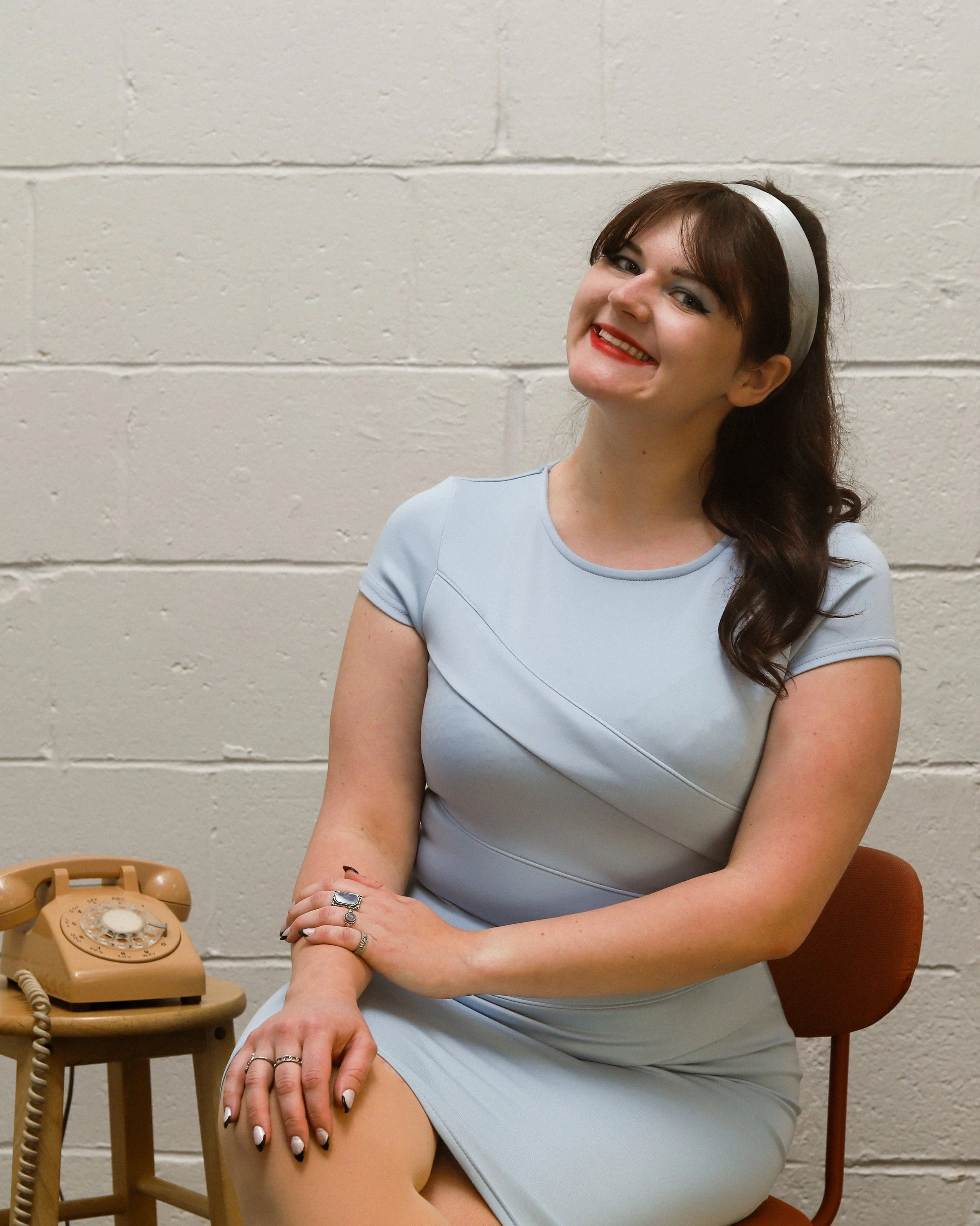 Cheyenne, a white woman, sits smiling at the camera. She has brown hair with a blue headband, which matches her dress. The background is white brick.