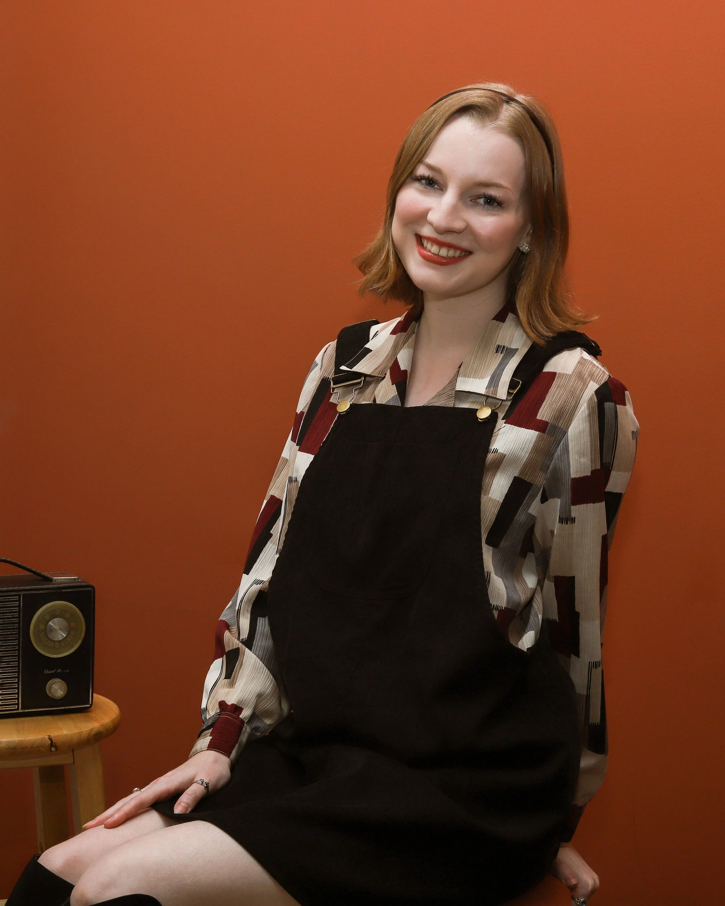 Emma, a white person, is sitting smiling at the camera. They have short blonde hair with a thin black headband. She is wearing a patterned blouse and a black overall dress. The background is terracotta.