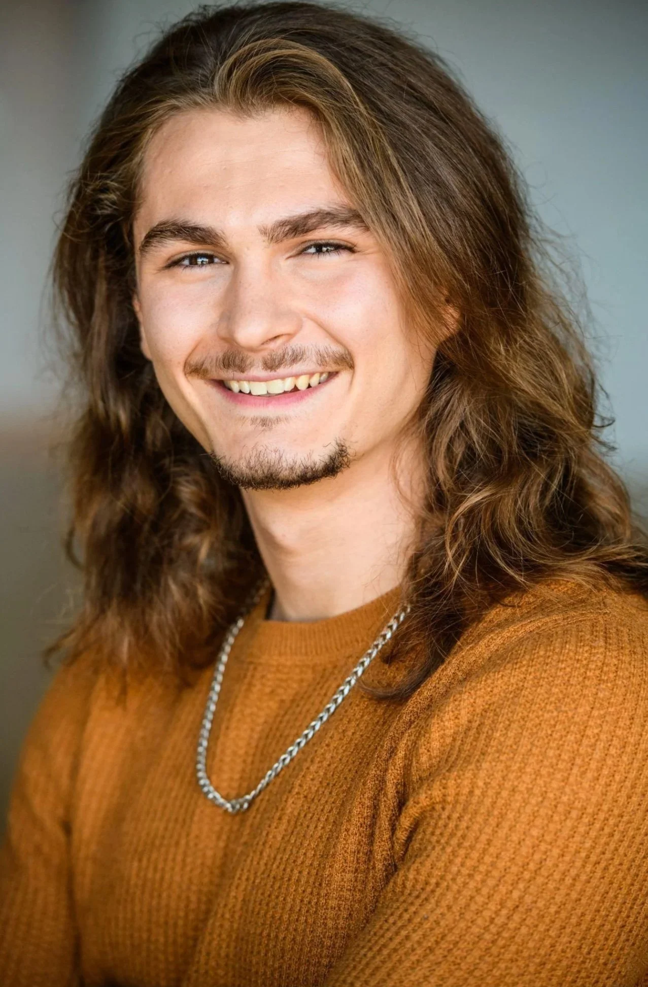 Blake, a white man, smiles at the camera. He has shoulder-length brown hair, and a goatee. He is wearing a pumpkin coloured sweater and a silver chain necklace.