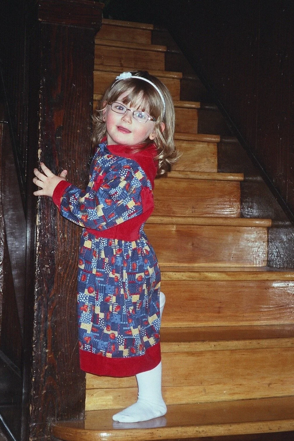 Little Emilia is smiling at the camera, standing on a staircase, hugging the wall. She is wearing a red and blue patterned dress, white stockings, a white headband, and glasses.