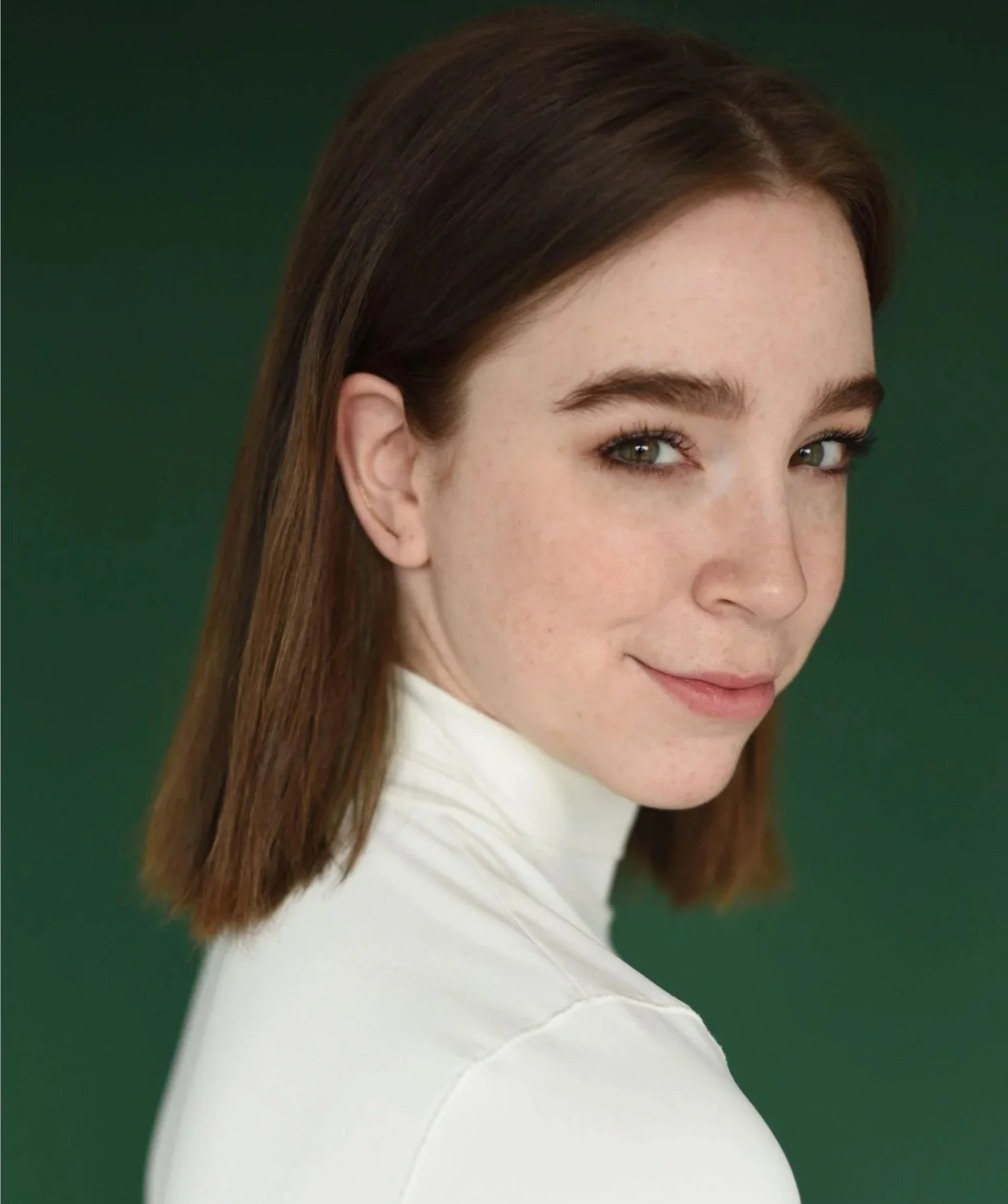 Madeleine, a nonbinary performer, is in front of a green background. They have a brown bob, and are smirking with mischief at the camera. They are waring a white turtleneck.