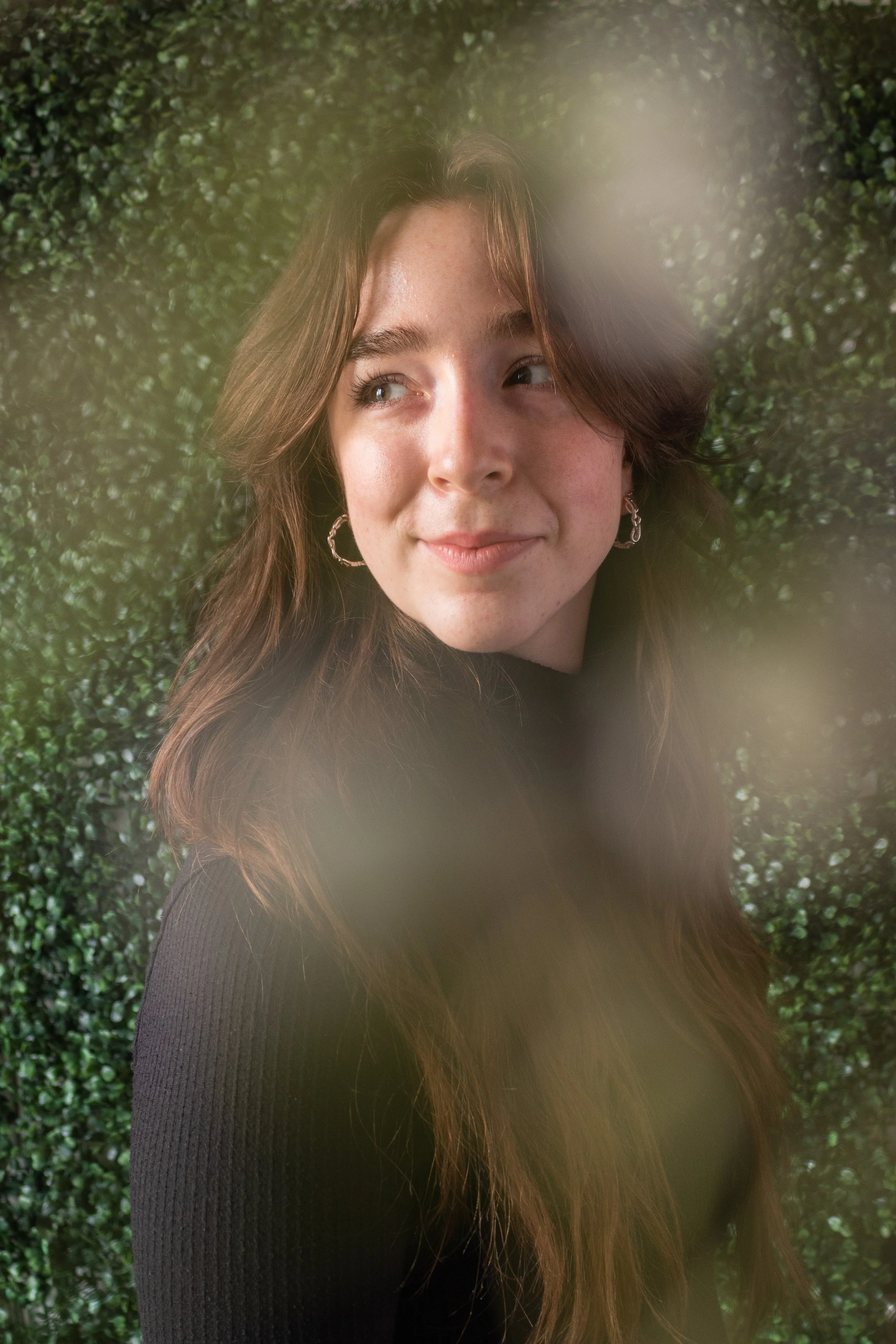Madeleine, a white person is smiling at something outside of frame. They have long brown hair. She is wearing a black turtleneck and gold earrings. Around the edge of frame are blurry white flowers, the background is green leaves.