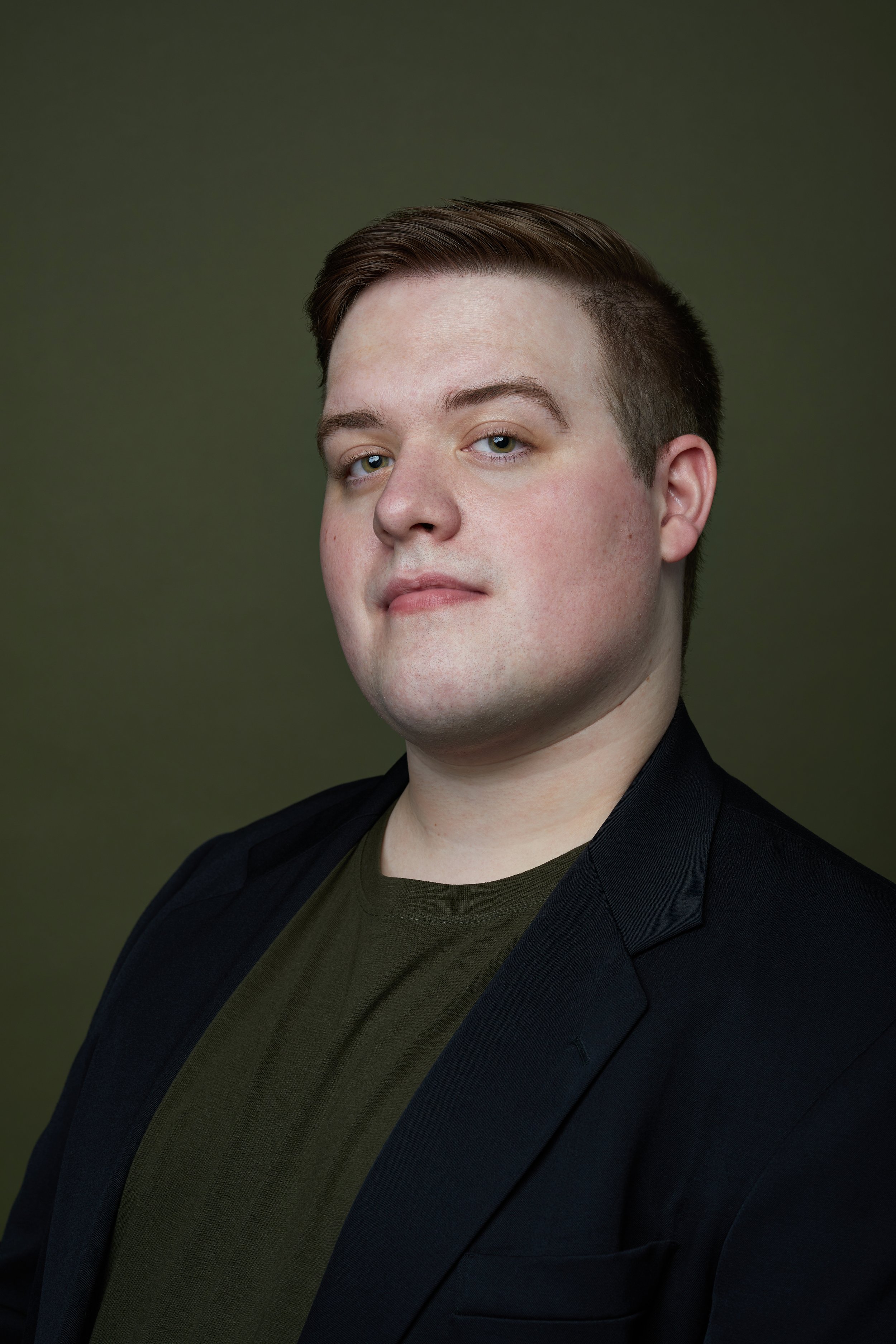 Aaron is a white man with short brown hair and green eyes. He is wearing a dark green shirt that matches the background, and a navy blazer.