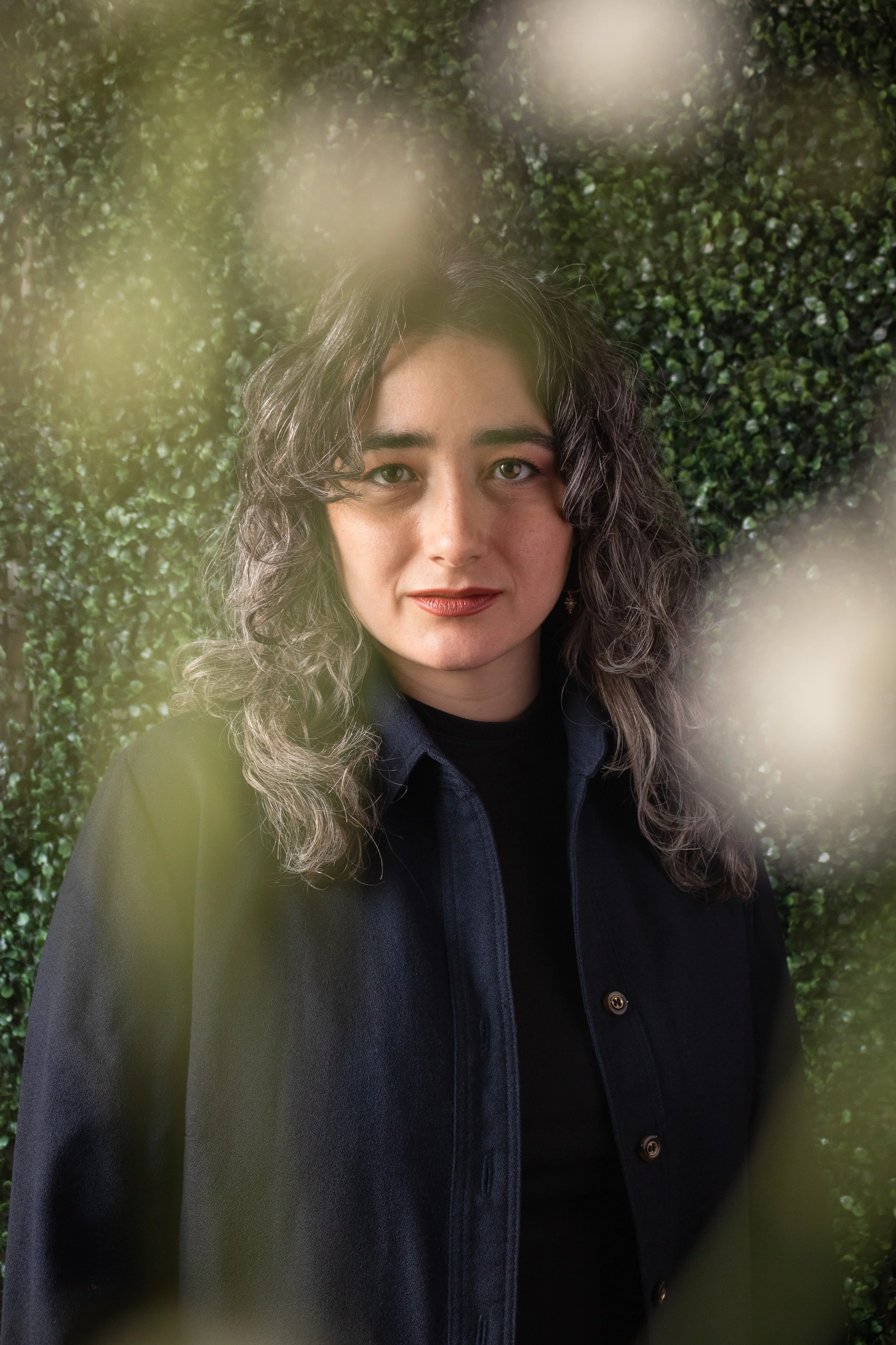 Carling, a white woman, looks determinedly at the camera. She has long curly salt and pepper hair, and is wearing a black button down with a turtleneck. Around the edge of frame are blurry white flowers, the background is green leaves.