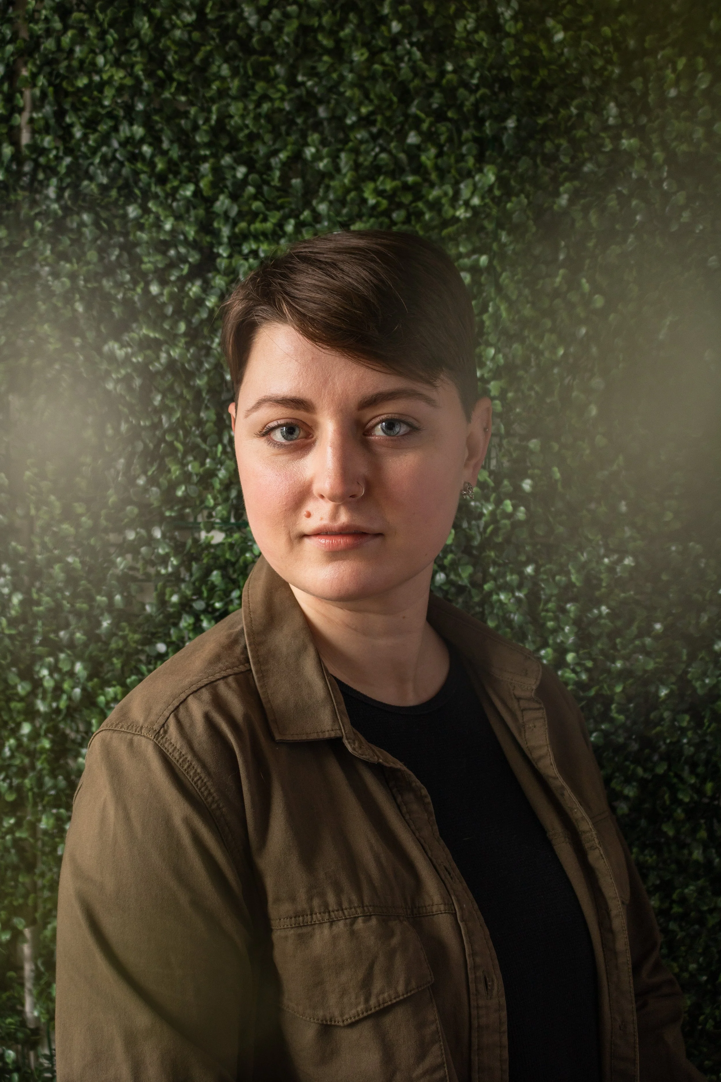 Hadley, a white person, is looking to the camera. They have short brown hair, and are wearing an olive green jacket over a black top. Around the edge of frame are blurry white flowers, the background is green leaves.
