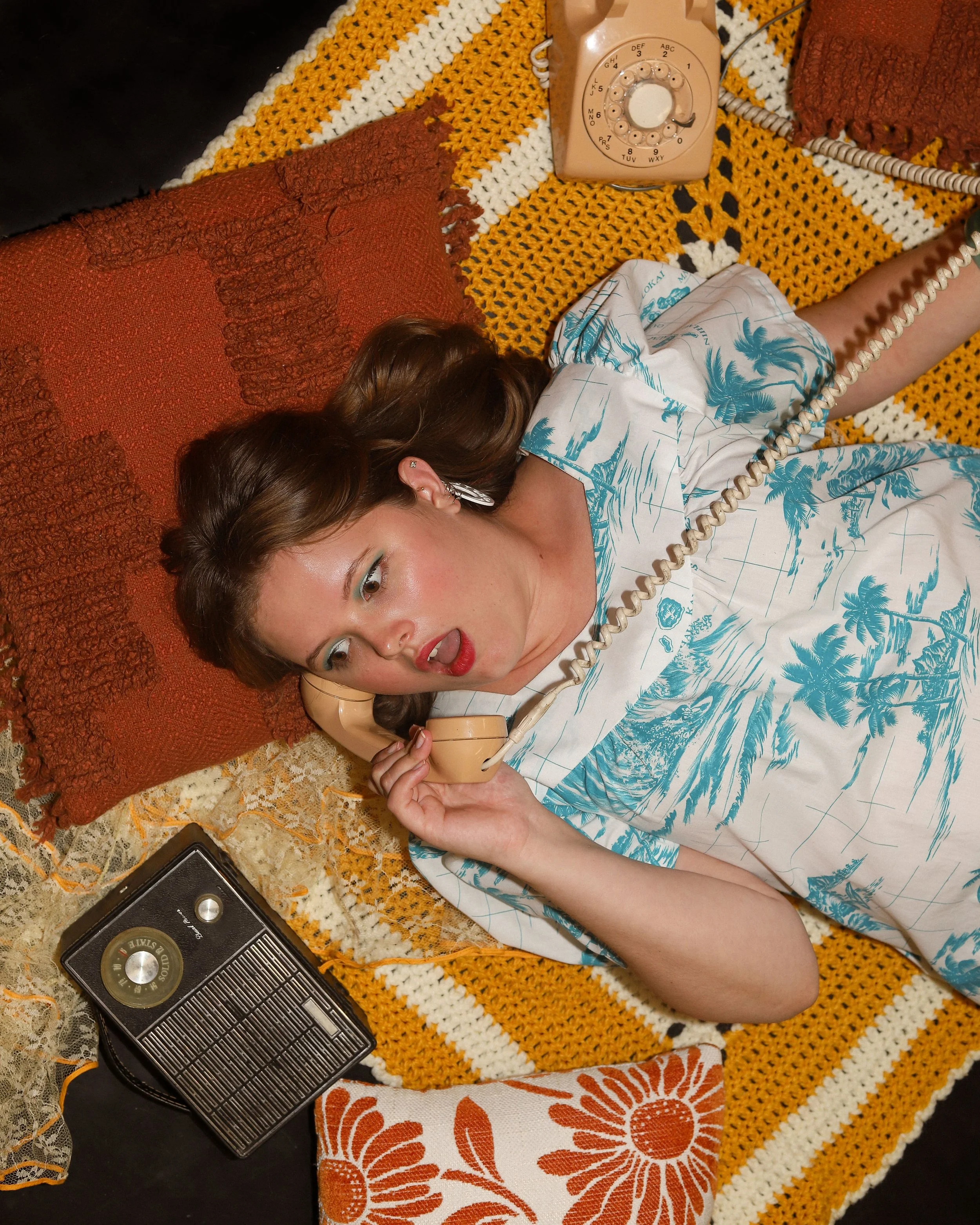 Meaghan, a white woman, lays against a collection of orange pillows and a white and yellow knit blanket. She is holding a rotary phone to her ear, and to her side is an old radio. Her brown hair is down and she has a surprised expression.