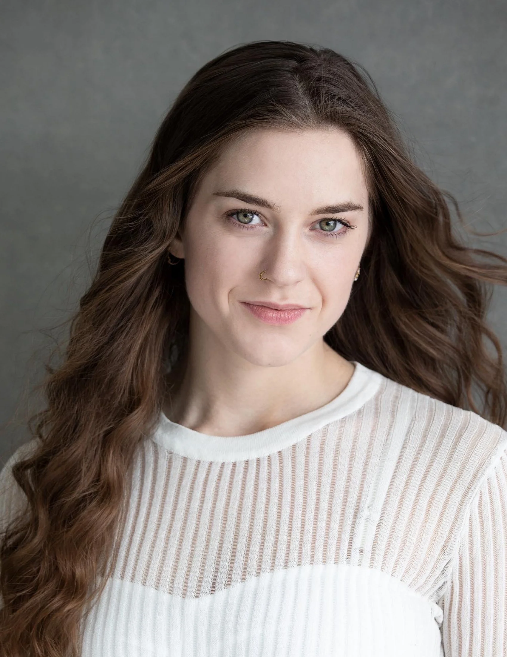 Enya is a white woman with long brown hair and blue eyes. She is smiling at the camera, and wearing a white sheer shirt. The background is grey.