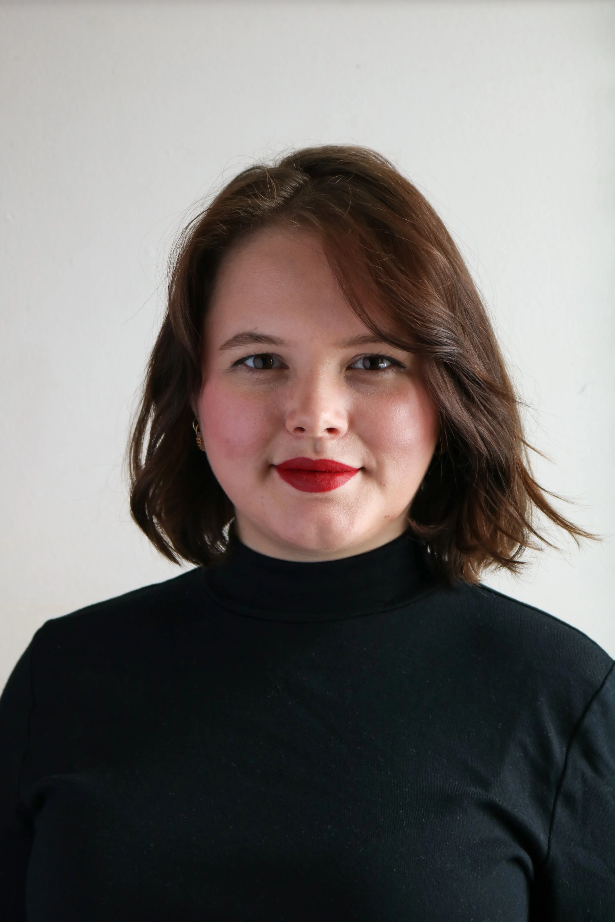 Meaghan, a white woman, smiles at the camera. She has a dark brown bob and is wearing red lipstick and a black turtleneck. The background is white.