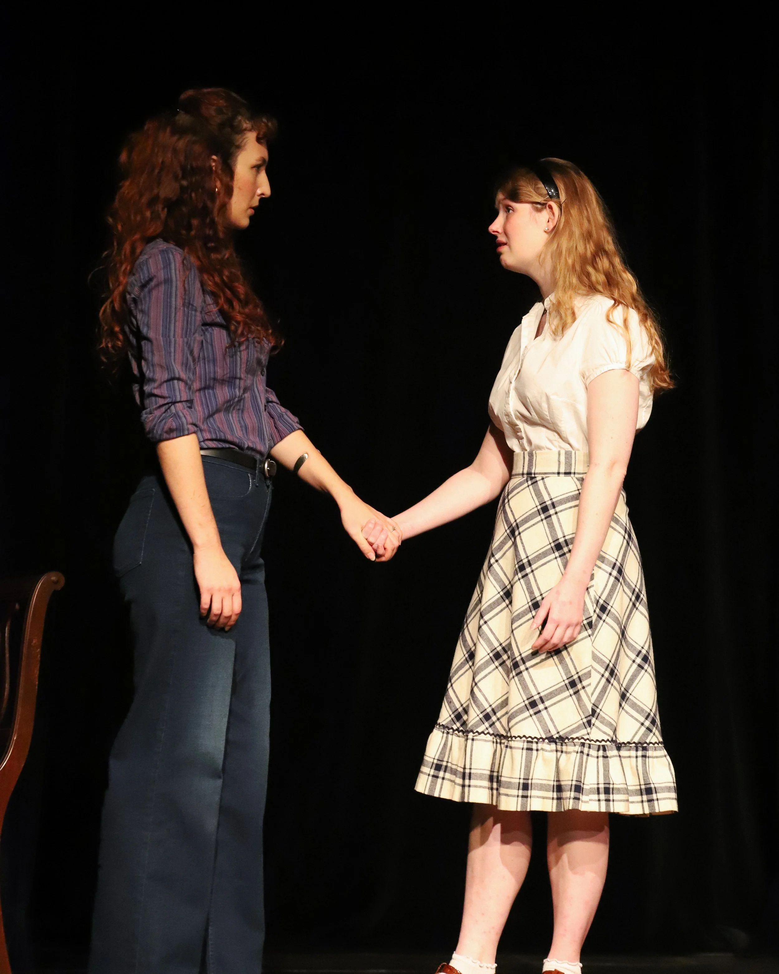 Two women stand facing each other, holding hands. They are dressed in clothing from the late 1960s. They are on stage, in front of a black backdrop.