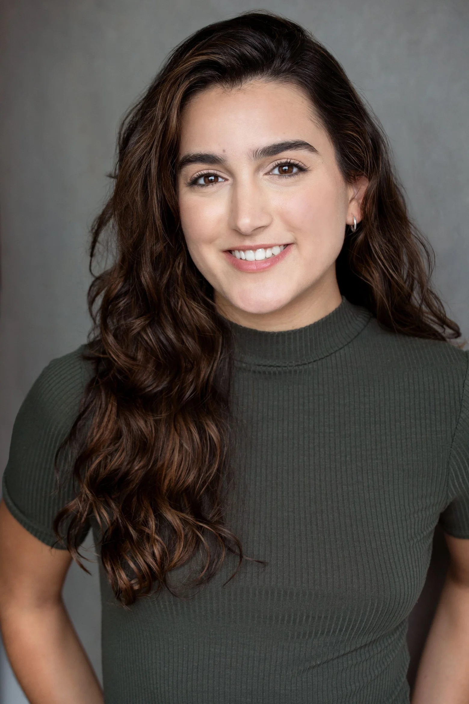 Freya is a Maltese Canadian actress with long brown hair. She is smiling at the camera, and wearing a grey mock neck shirt. She stands in front of a grey background.