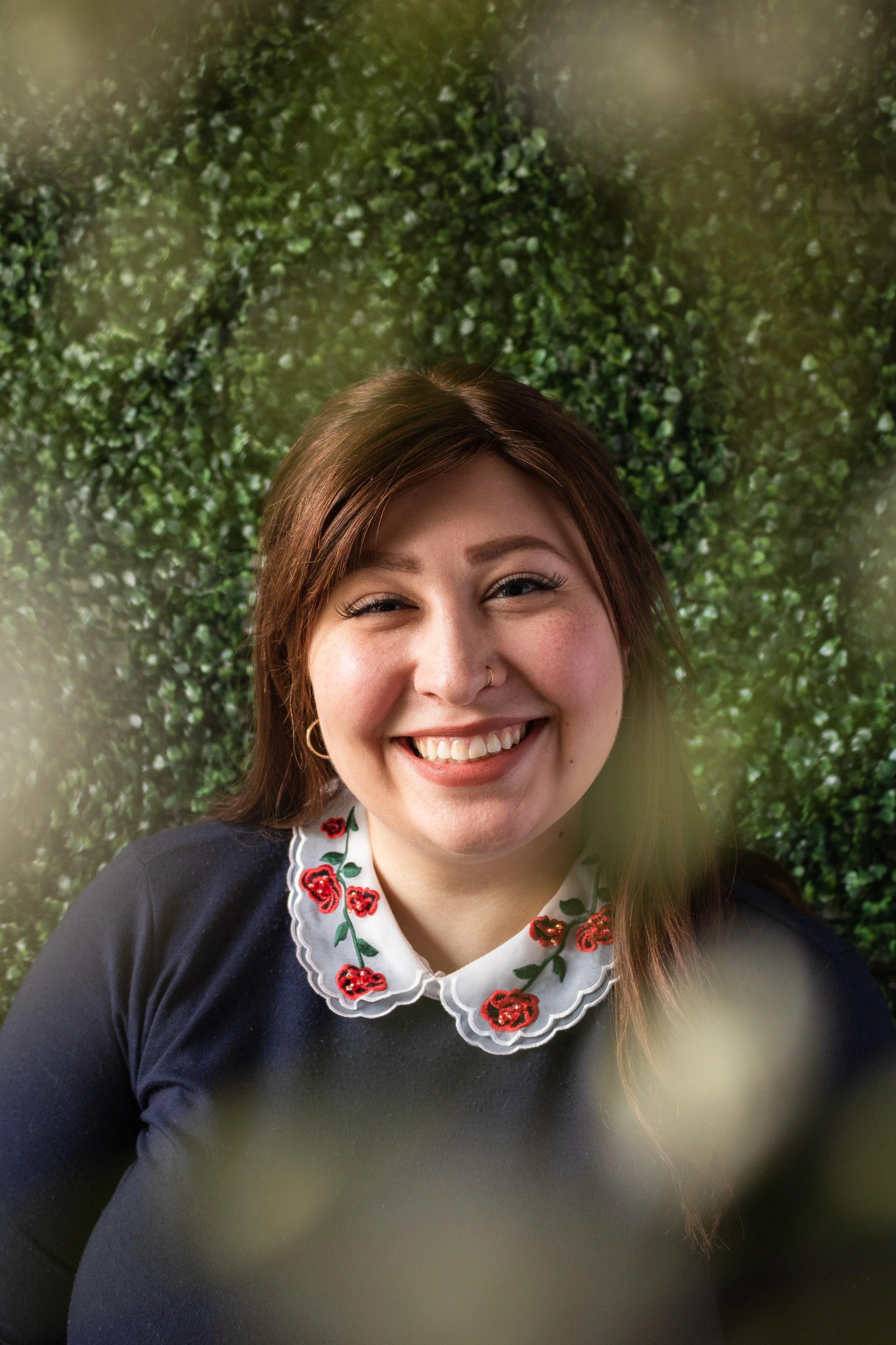 Josephine, a white woman, is smiling at the camera. She has long brown hair, and is wearing a navy shirt with a white peter pan collar, that has roses embroidered. Around the edge of frame are blurry white flowers, the background is green leaves